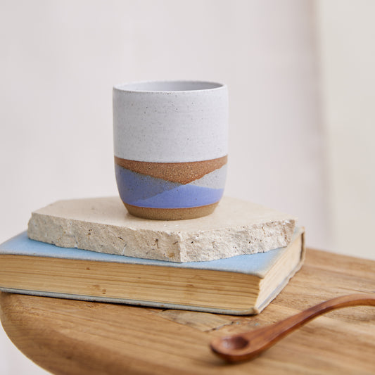 A handmade ceramic tumbler sitting on a book and side table. The handmade ceramic tumbler is glazed in a white, violet and grey glaze.