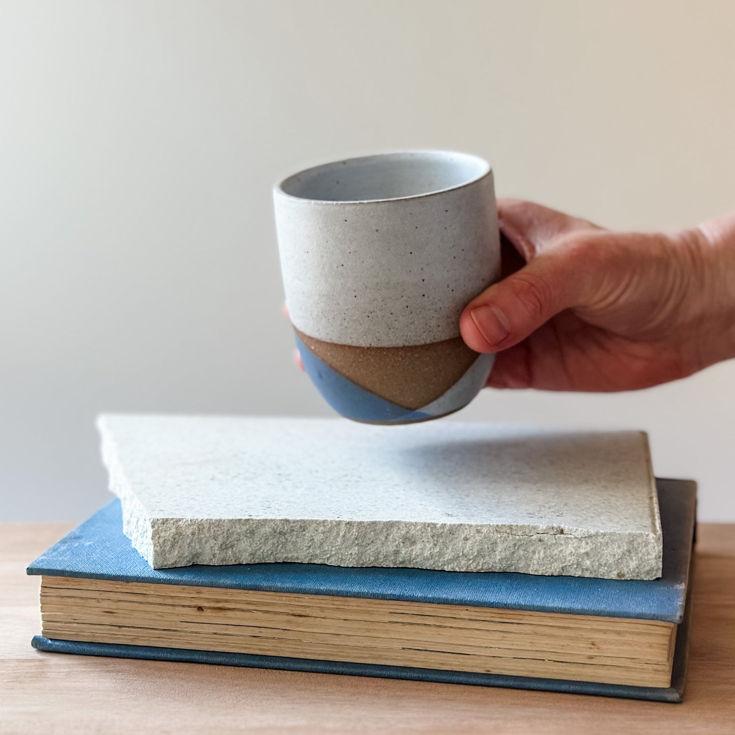 Hand holding a ceramic cup over two stacked books on a wooden surface.