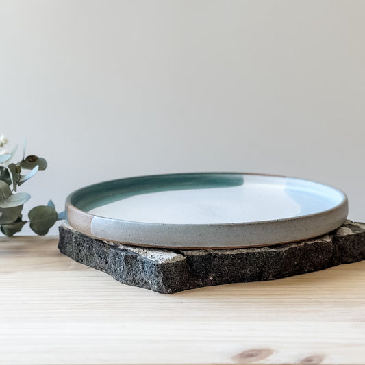 The small Tidal Handmade Ceramic Plate in a white, green and blue-grey glaze. The plate sits on a tile and a wooden table.
