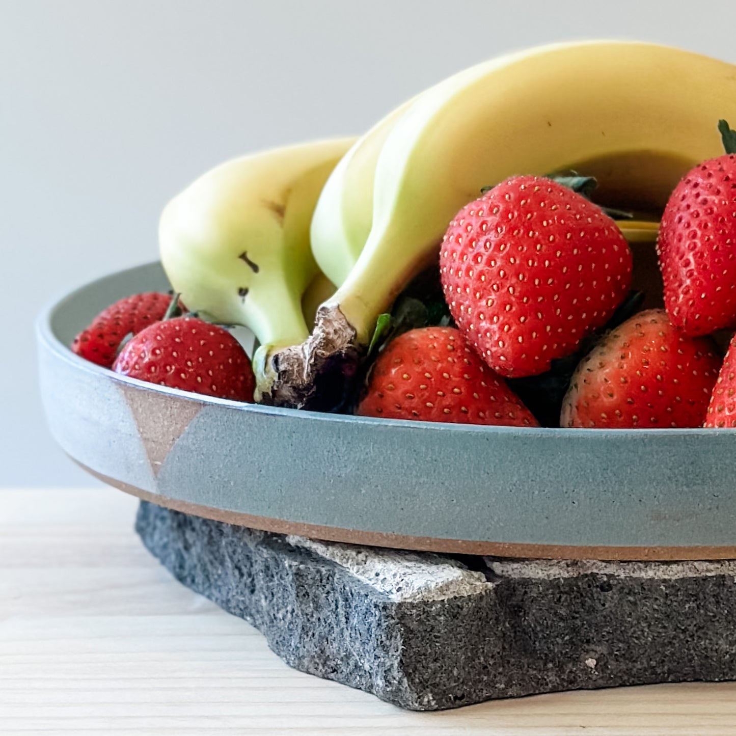A close-up view of the large Tidal Handmade Ceramic Platter sitting on a tile and a wooden table. The handmade platter is green, blue-grey and white and holds bananas and strawberries.