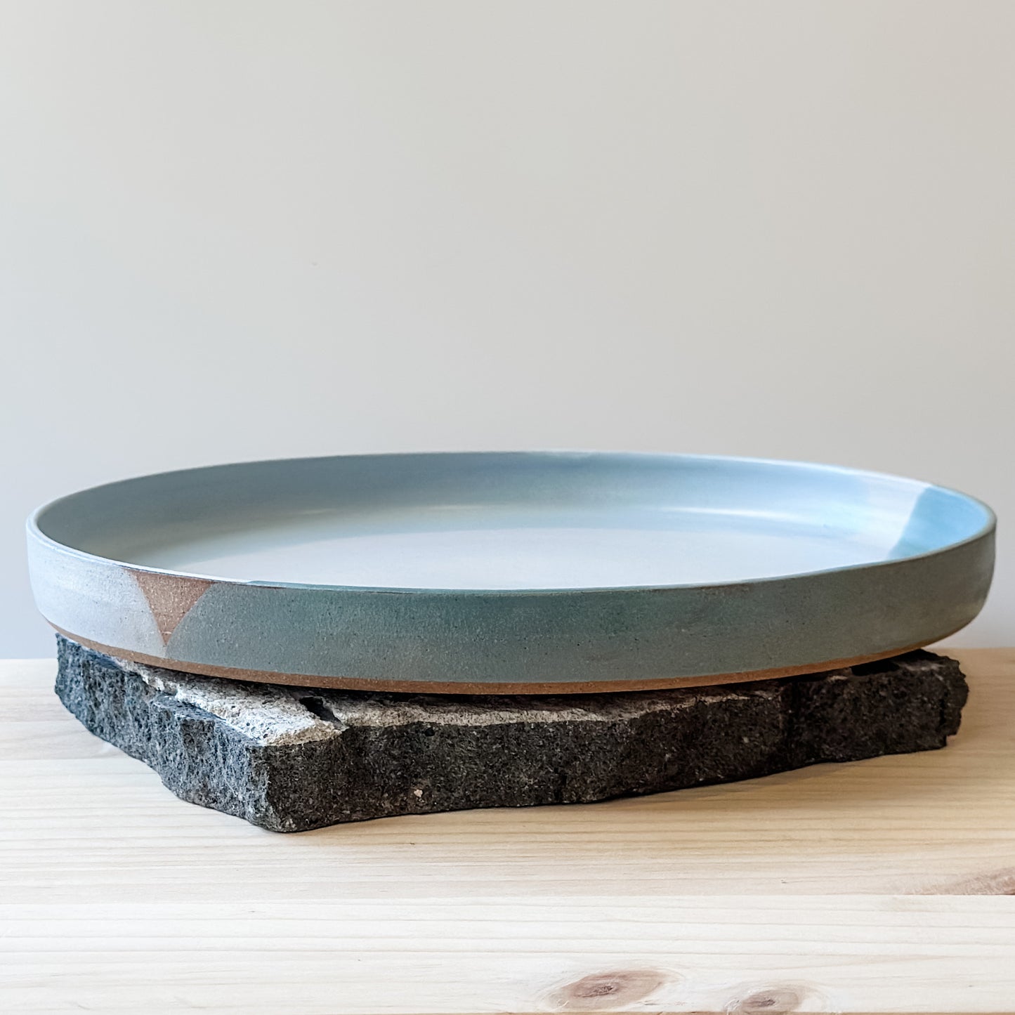 A front view of the large Tidal Handmade Ceramic Platter sitting on a tile and a wooden table. The handmade platter is green, blue-grey and white.