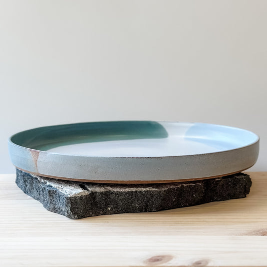 A large Tidal Handmade Ceramic Platter sits on a tile and a wooden table. The handmade platter is green, blue-grey and white.