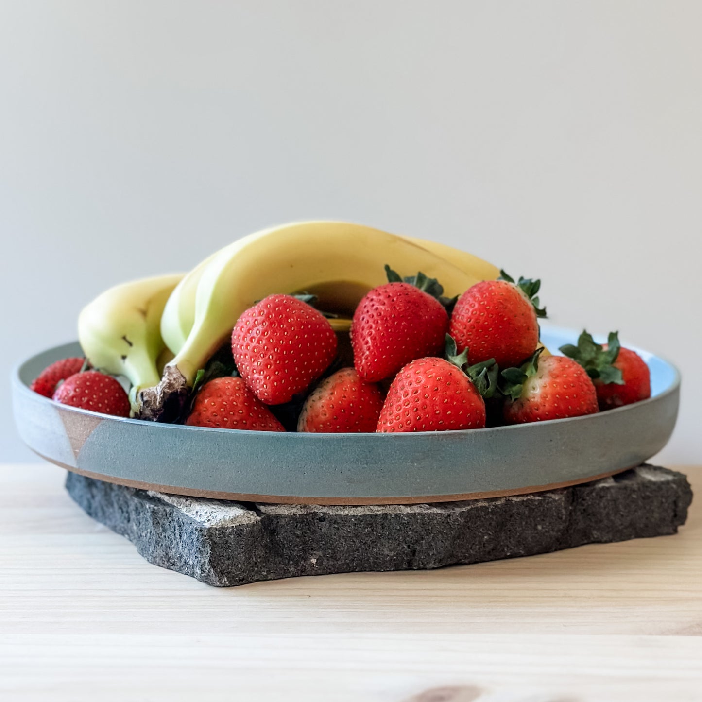 A side view of the large Tidal Handmade Ceramic Platter sitting on a tile and a wooden table. The handmade platter is green, blue-grey and white and holds bananas and strawberries.