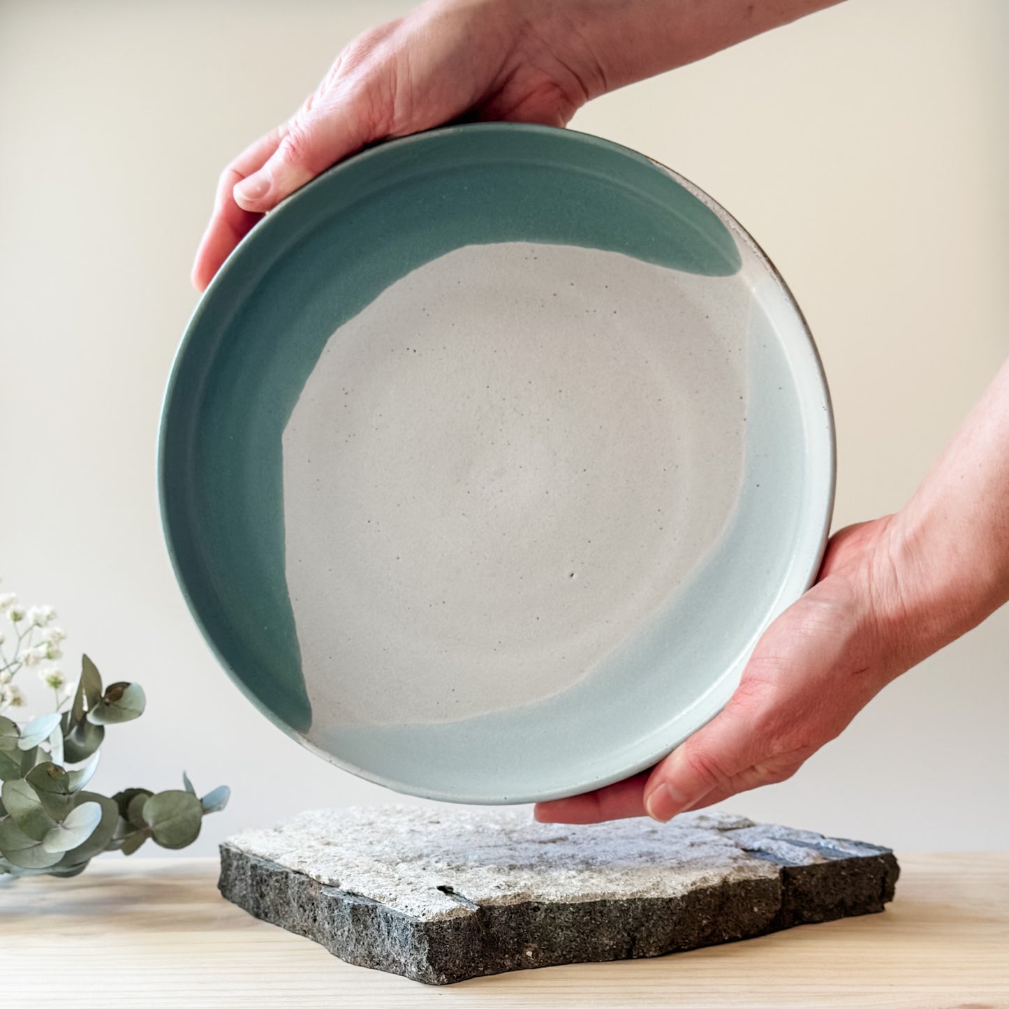 A view of the inside of the medium-sized Tidal Handmade Ceramic Platter on a tile and a wooden table. The platter is glazed in green, blue-grey and white. The platter is being held by two hands.