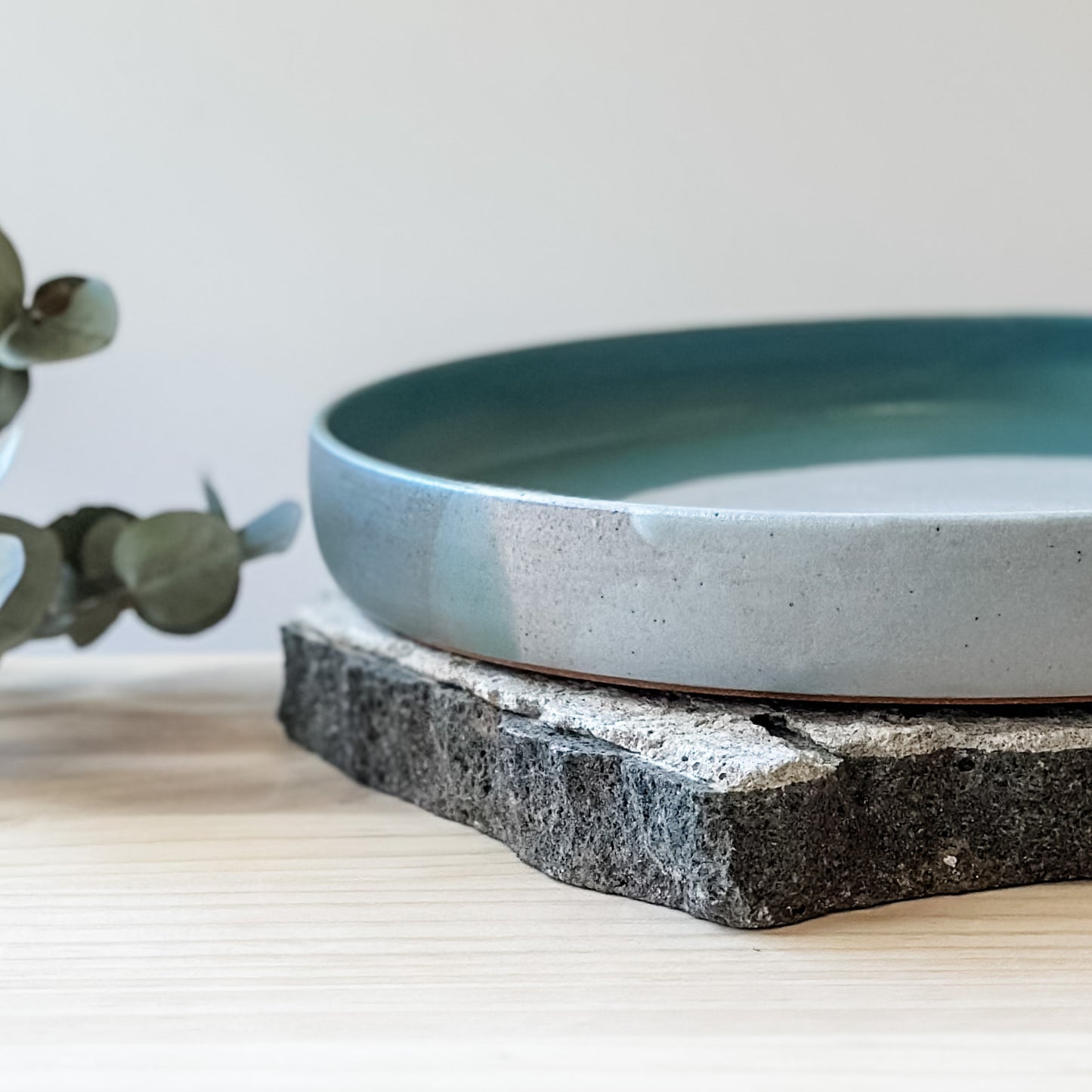 A close-up view of the medium-sized Tidal Handmade Ceramic Platter on a tile and a wooden table. The platter is glazed in green, blue-grey and white.