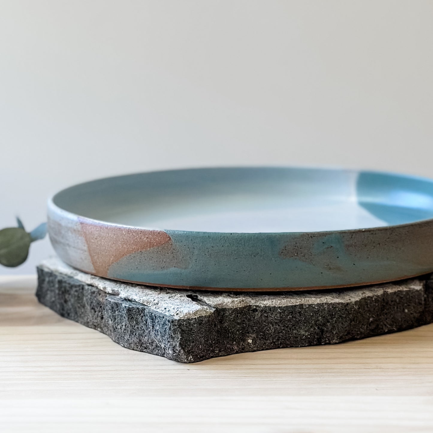 A close-up view of the medium-sized Tidal Handmade Ceramic Platter on a tile and a wooden table. The platter is glazed in green, blue-grey and white.