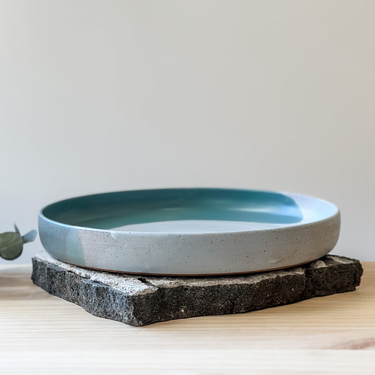 A front view of the medium-sized Tidal Handmade Ceramic Platter on a tile and a wooden table. The platter is glazed in green, blue-grey and white.