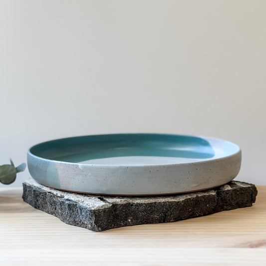 A front view of the medium-sized Tidal Handmade Ceramic Platter on a tile and a wooden table. The platter is glazed in green, blue-grey and white.