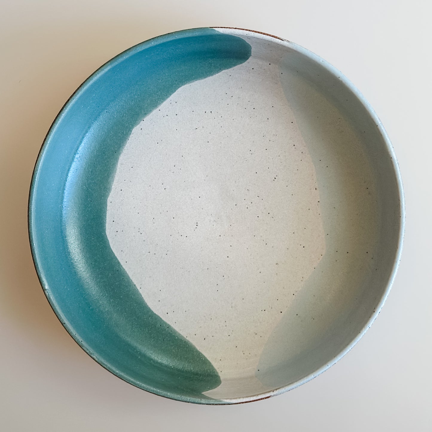 An aerial view of the Tidal Handmade Ceramic Serving Bowl on a cream background. The handmade bowl is glazed green, blue-grey and white.