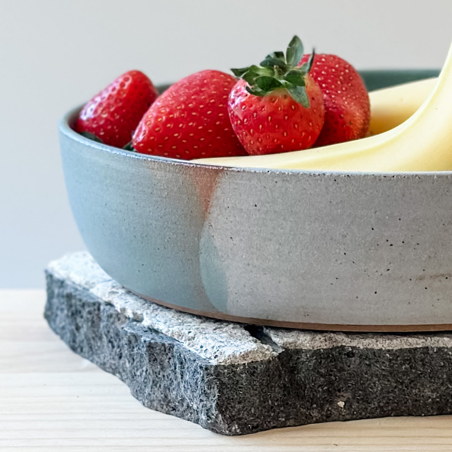 A close-up view of the Tidal Handmade Ceramic Serving Bowl. The handmade bowl holds strawberries and bananas and sits on a tile and a wooden table.