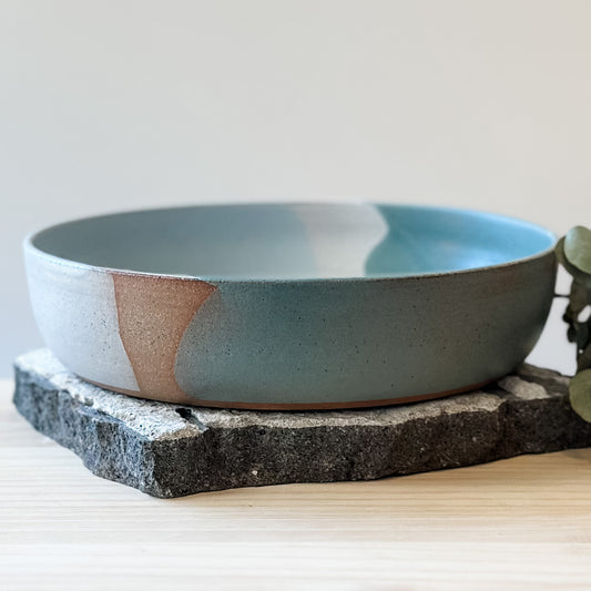 A side view of the Tidal Handmade Ceramic Serving Bowl. The handmade bowl sits on a tile and a wooden table.