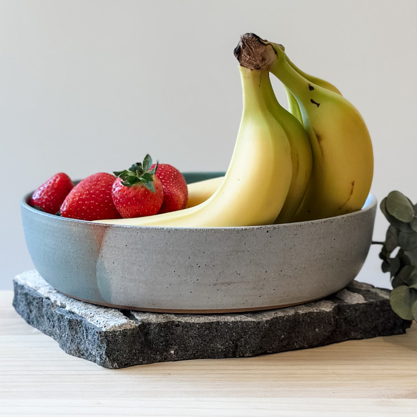 A side view of the Tidal Handmade Ceramic Serving Bowl with strawberries and bananas. The handmade bowl is glazed green, blue-grey and white and sits on a tile and a wooden table.