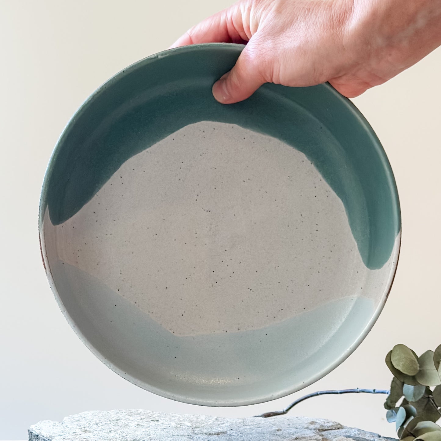 An interior view of the Tidal Handmade Ceramic Serving Bowl. The handmade bowl is glazed green, blue-grey and white and is held with one hand above a tile.