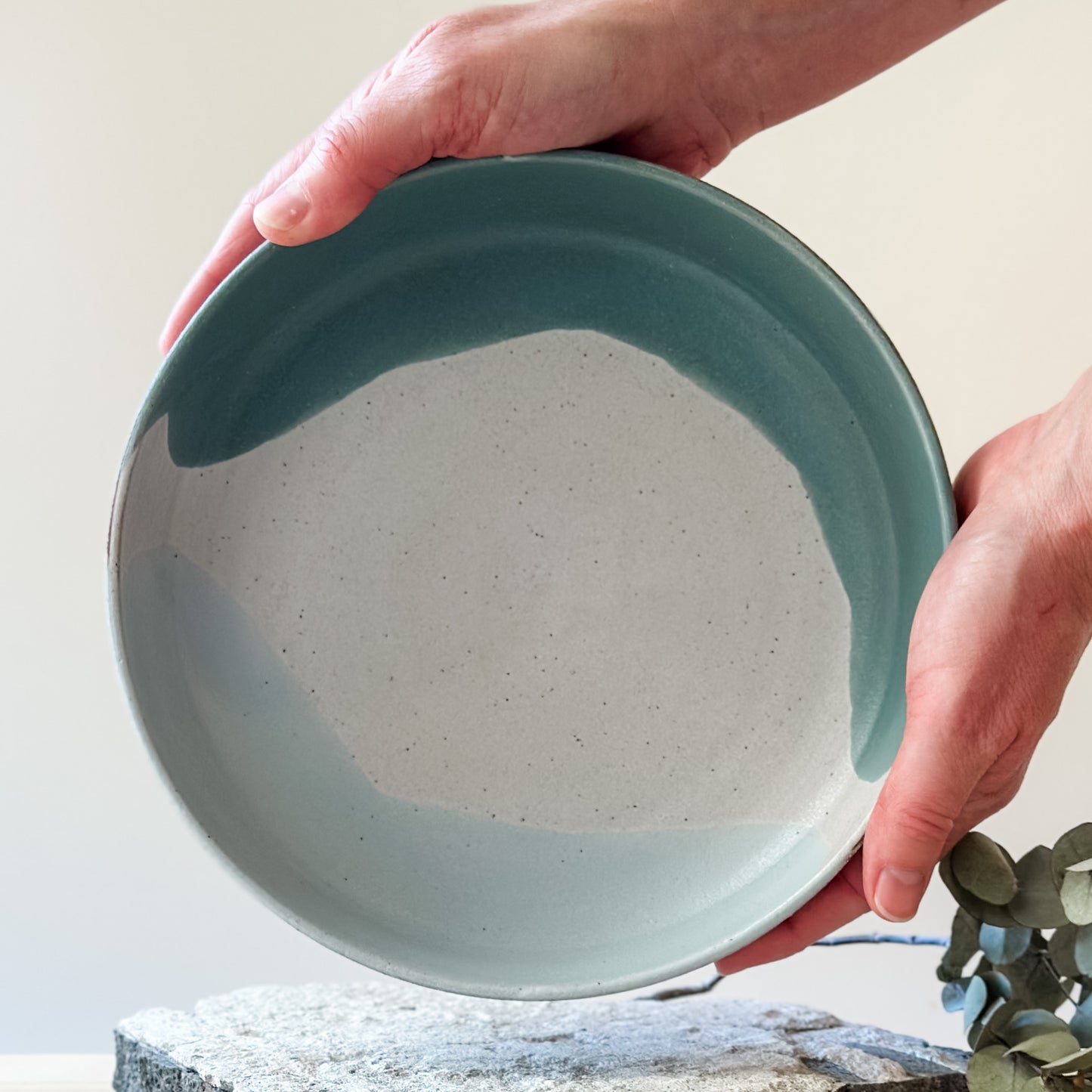 An interior view of the Tidal Handmade Ceramic Serving Bowl. The handmade bowl is glazed green, blue-grey and white and is held with two hands above a tile.