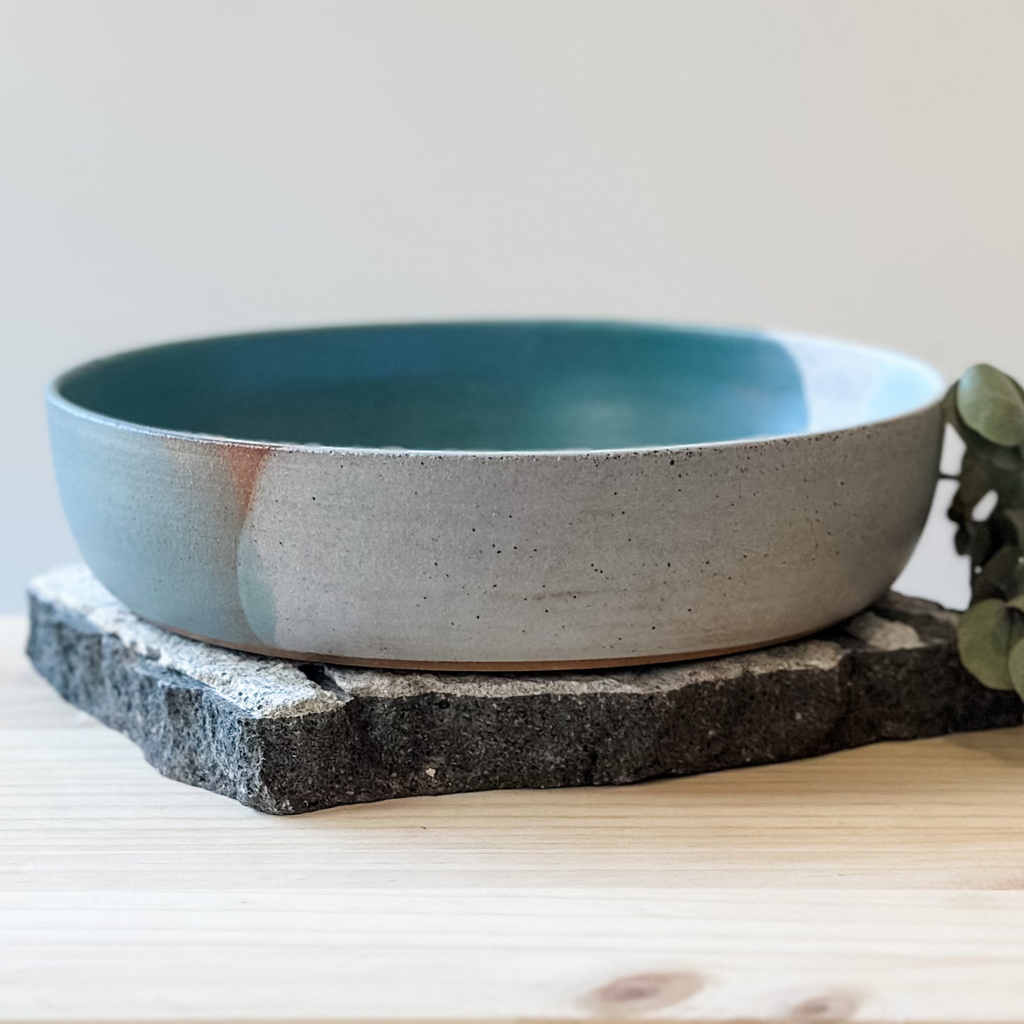 A side view of the Tidal Handmade Ceramic Serving Bowl. The handmade bowl is glazed green, blue-grey and white and sits on a tile and a wooden table.