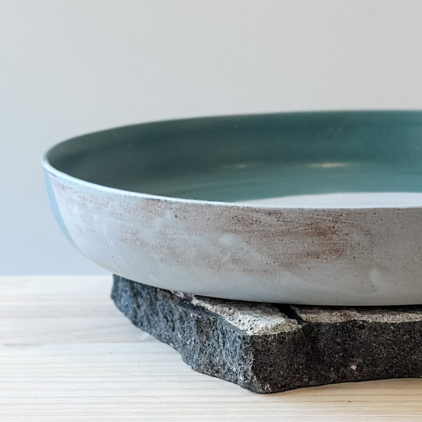 A close-up view of the side of the large Tidal Handmade Ceramic Serving Dish. The dish is green, white and blue-grey and sits on a tile and a wooden table.