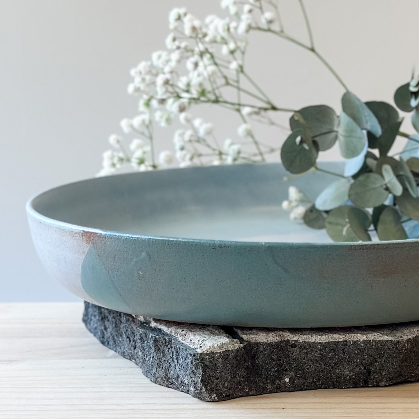 A close-up view of the large Tidal Handmade Ceramic Serving Dish. The handmade dish holds baby's breath flowers and gum leaves. The dish is green, white and blue-grey and sits on a tile and a wooden table.