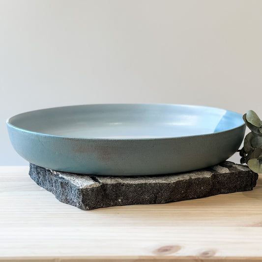 The large Tidal Handmade Ceramic Serving Dish sits on a tile and a wooden table. The handmade dish is green, white and blue-grey.