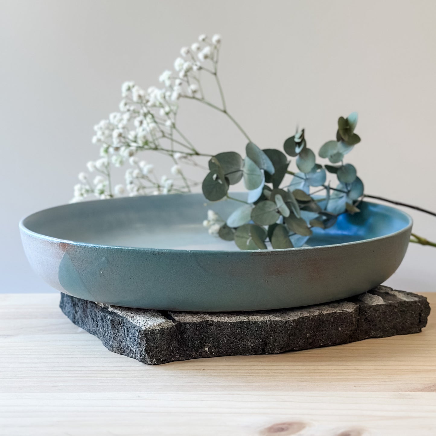 A view of the side of the large Tidal Handmade Ceramic Serving Dish. The handmade dish holds baby's breath flowers and gum leaves and is glazed green, white and blue-grey.