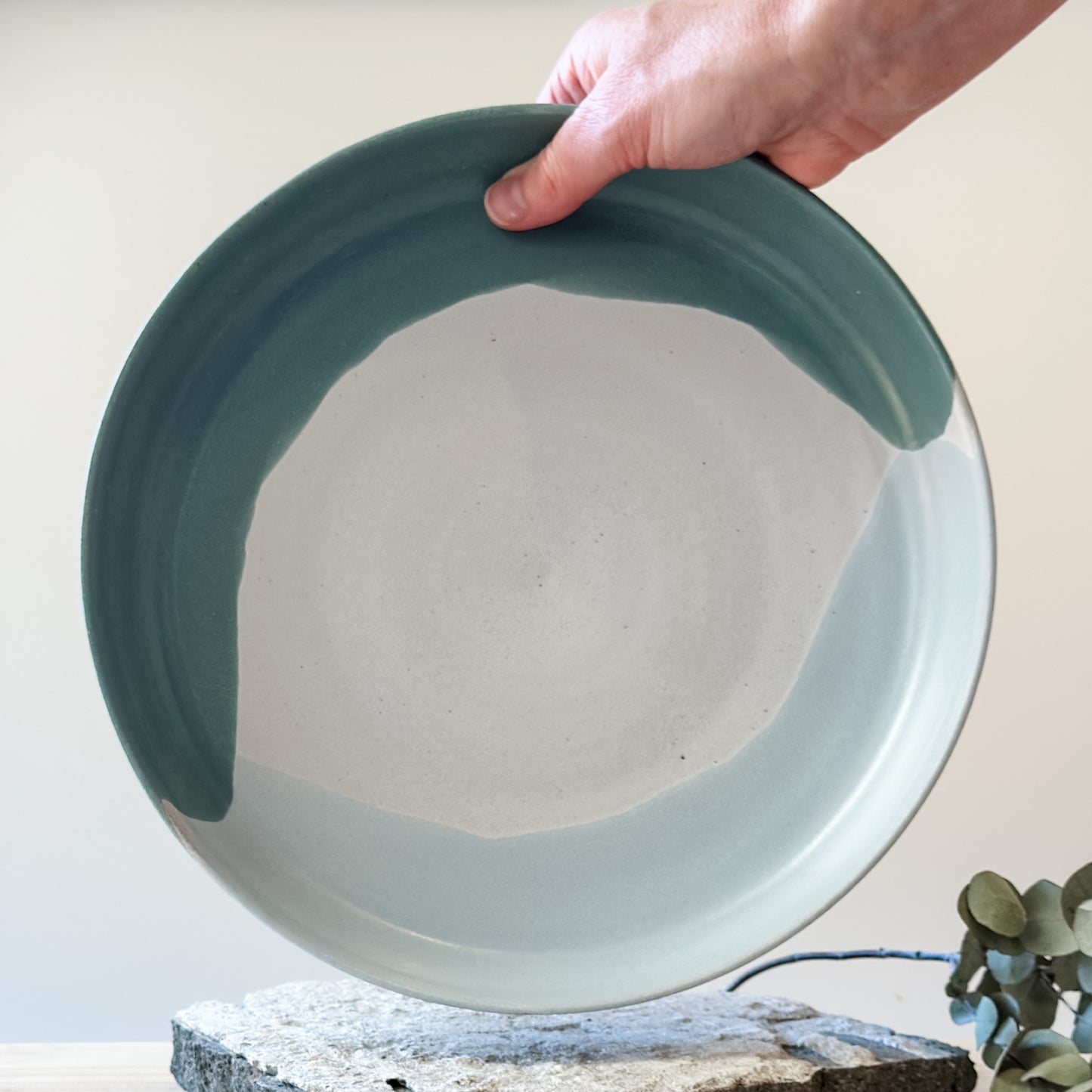 A view of the inside of the large Tidal Handmade Ceramic Serving Dish. The handmade dish is held with one hand above a tile and is glazed green, white and blue-grey.