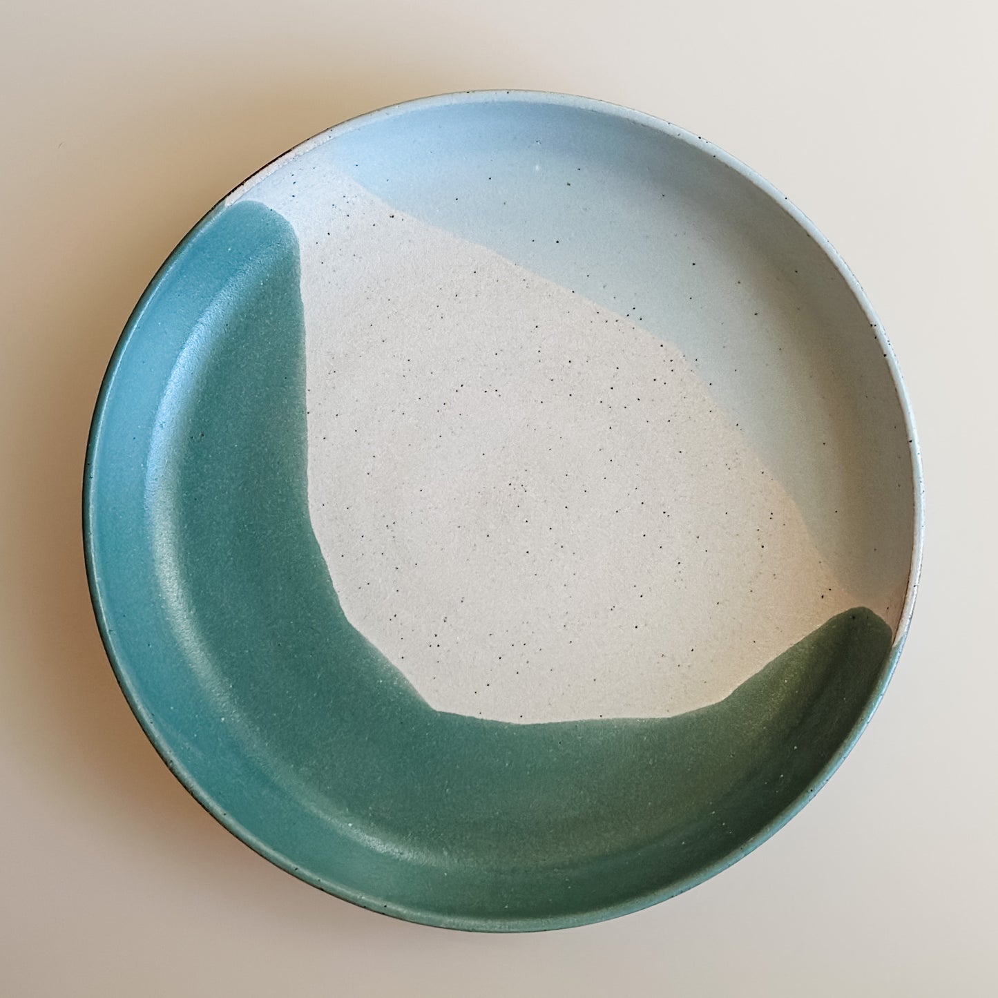 An aerial view of the medium Tidal Handmade Ceramic Dish in a green, white and blue-grey glaze. The ceramic dish sits on a cream background.