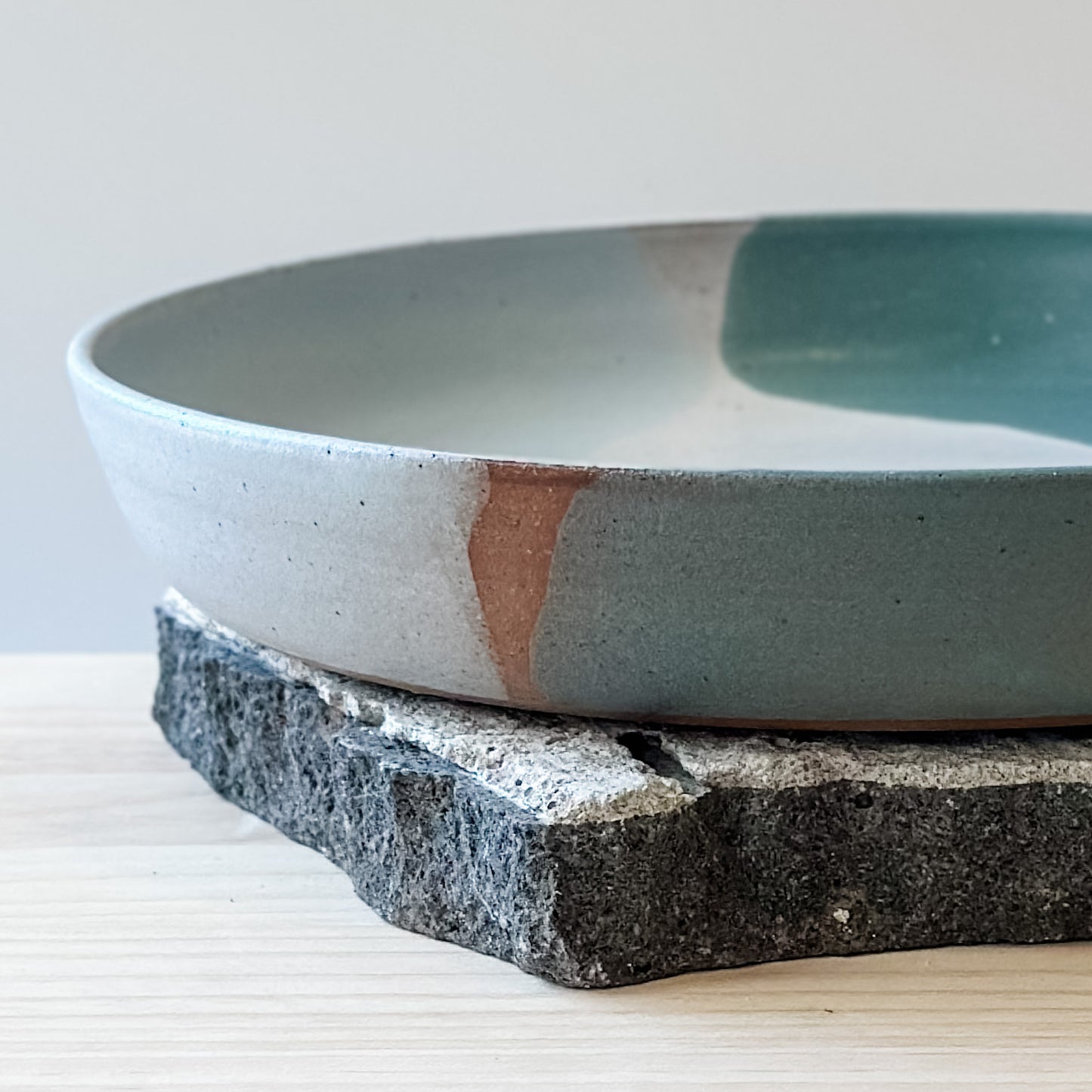 A close-up view of the medium Tidal Handmade Ceramic Dish in a green, white and blue-grey glaze. The ceramic dish sits on a tile and a wooden table.