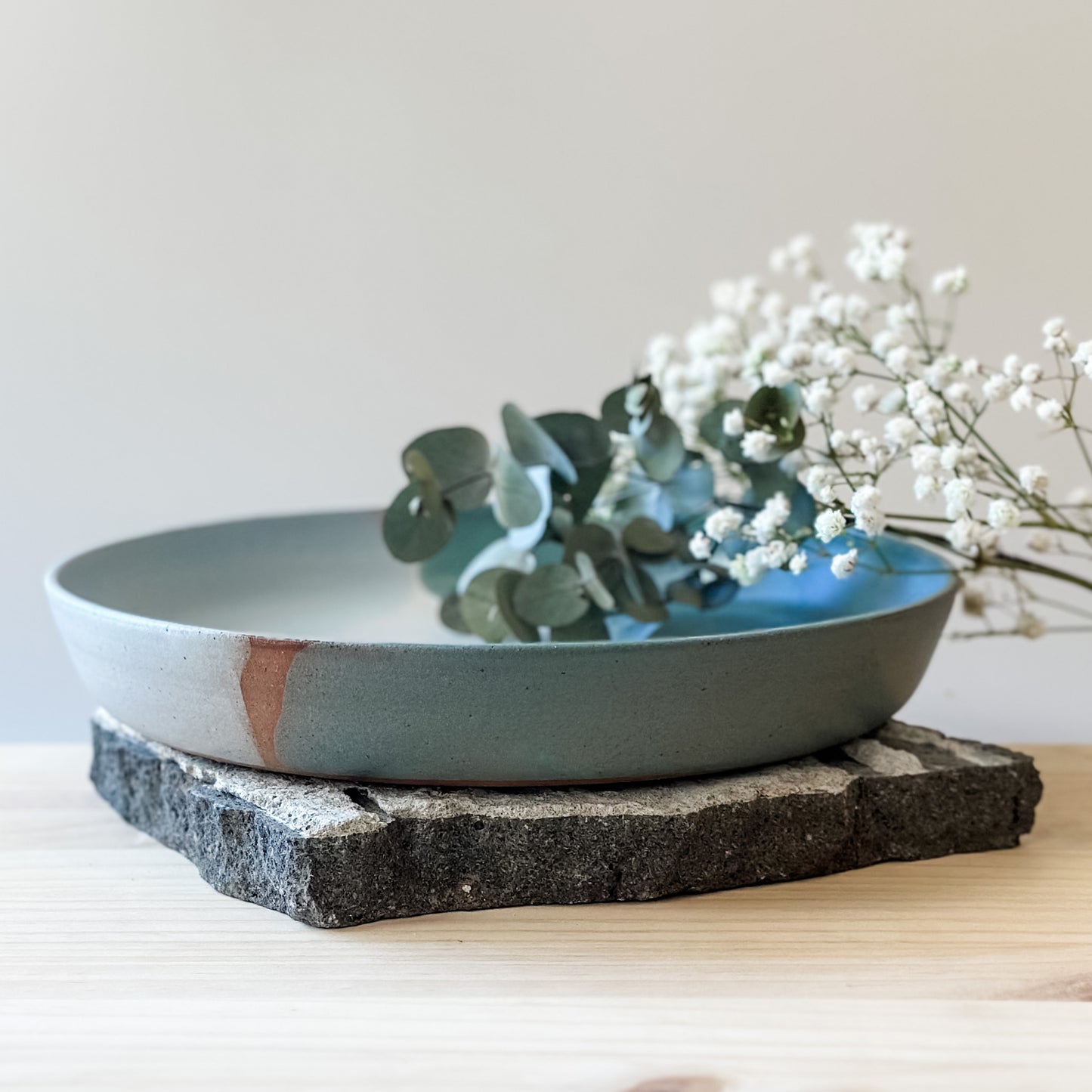 A front view of the medium Tidal Handmade Ceramic Dish in a green, white and blue-grey glaze. The ceramic dish hold flowers and sits on a tile and a wooden table.