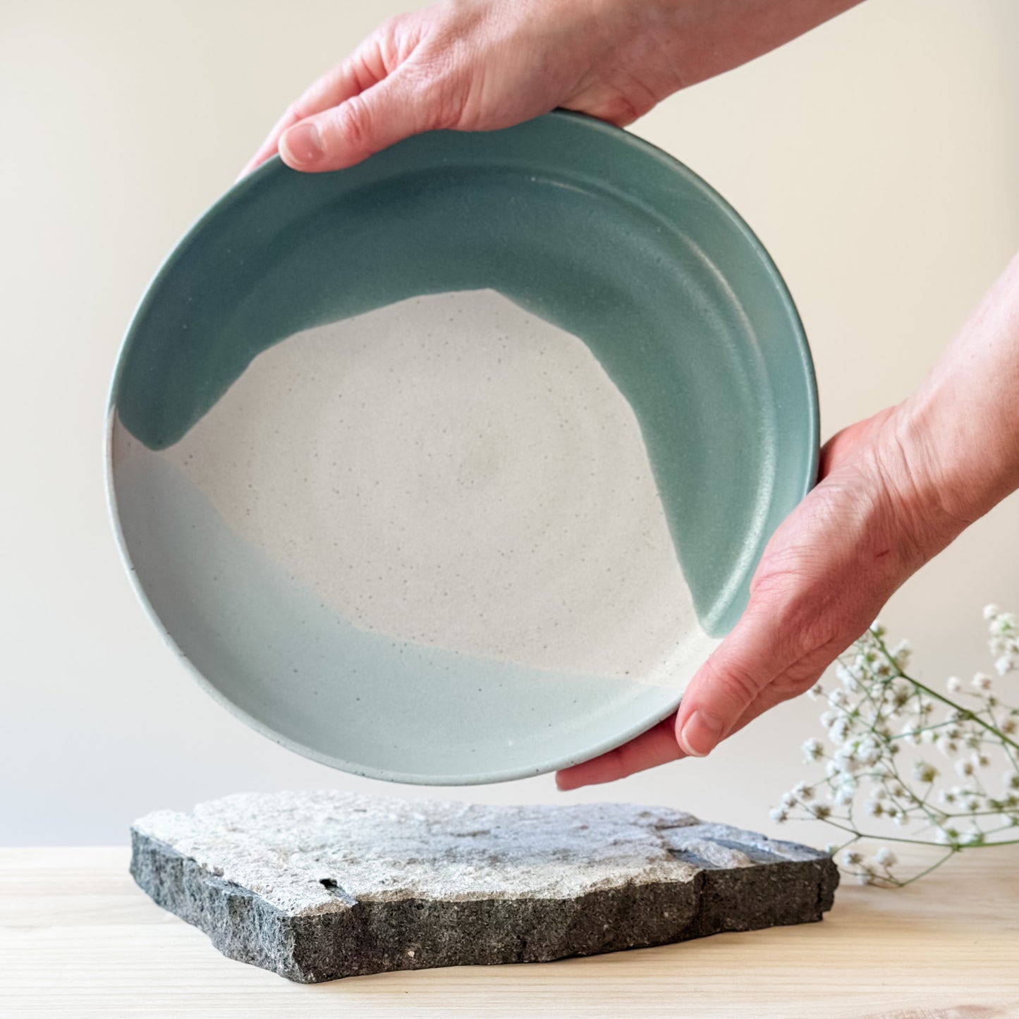 An interior view of the medium Tidal Handmade Ceramic Dish in a green, white and blue-grey glaze. The ceramic dish is held by two hands over a tile and a wooden table.