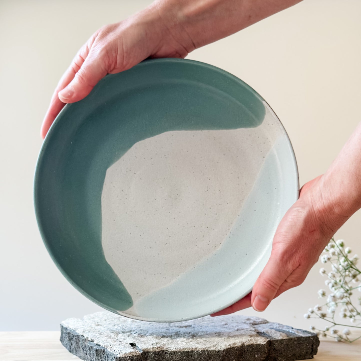An interior view of the medium Tidal Handmade Ceramic Dish in a green, white and blue-grey glaze. The ceramic dish is held by two hands over a tile and a wooden table.