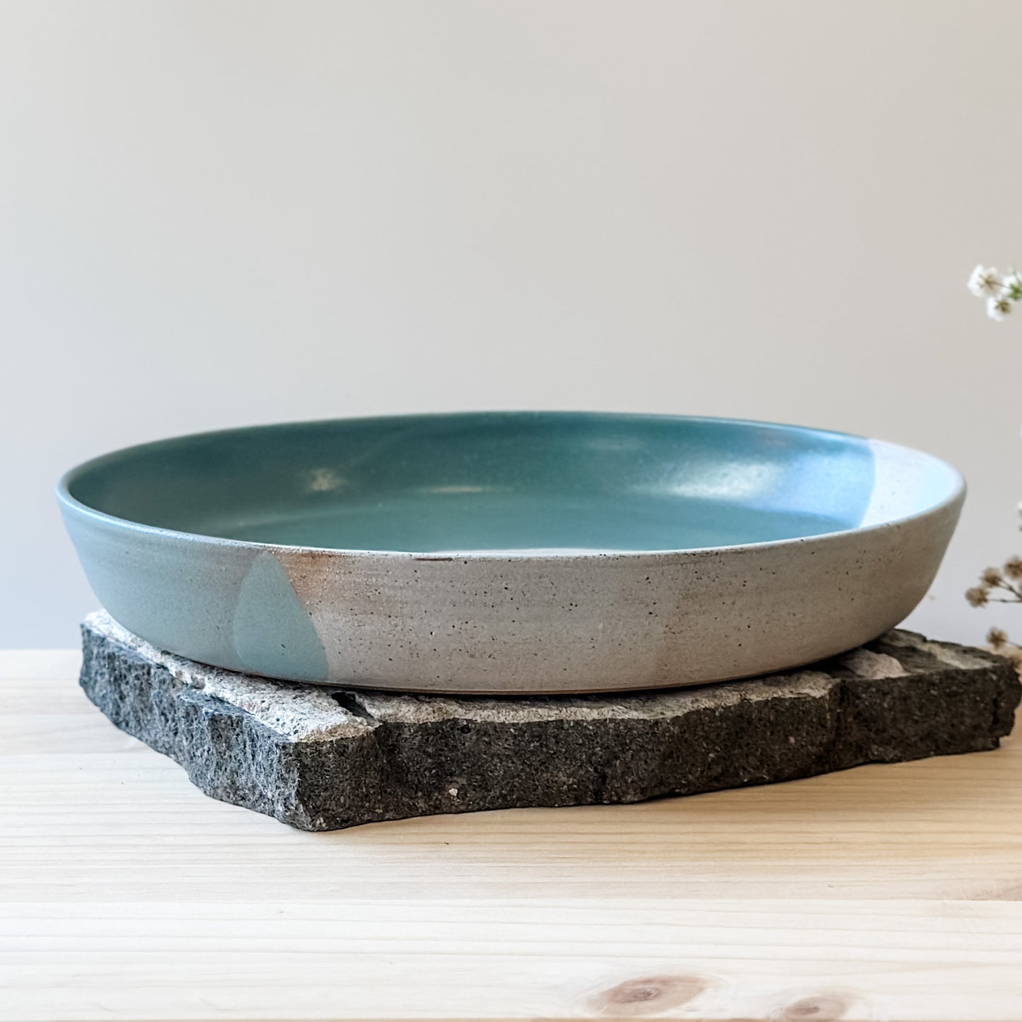 A front view of the medium Tidal Handmade Ceramic Dish in a green, white and blue-grey glaze. The ceramic dish sits on a tile and a wooden table.