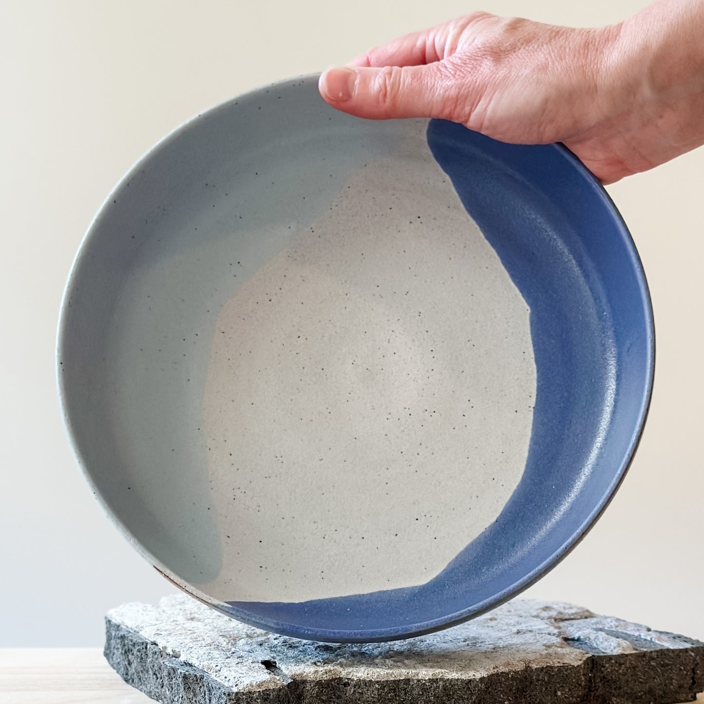 An interior view of the Tidal Handmade Ceramic Dish in a violet purple and blue-grey glaze. The handmade dish is held by one hand above a tile and a wooden table.