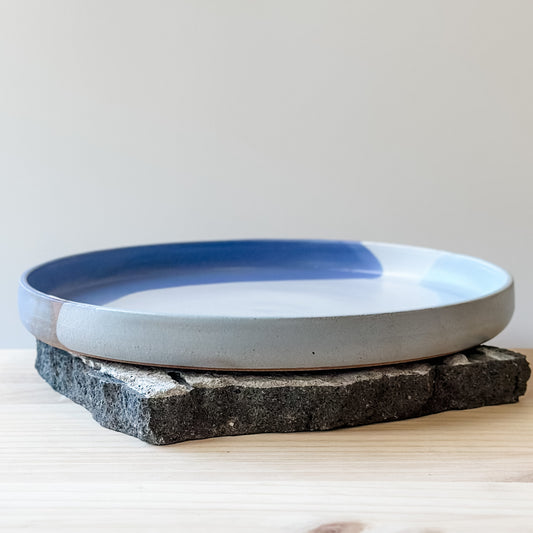 A front view of the medium Tidal Handmade Ceramic Platter in violet, blue-grey and white glaze. The platter sits on a tile and a wooden table.