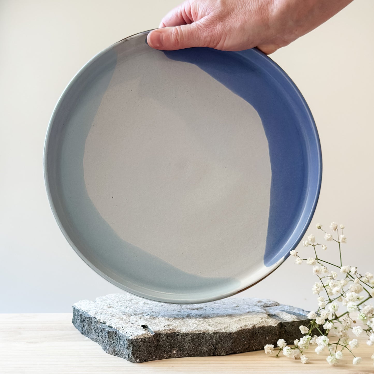 An interior view of the medium Tidal Handmade Ceramic Platter in violet, blue-grey and white glaze. The platter is held by one hand above a tile and a wooden table.