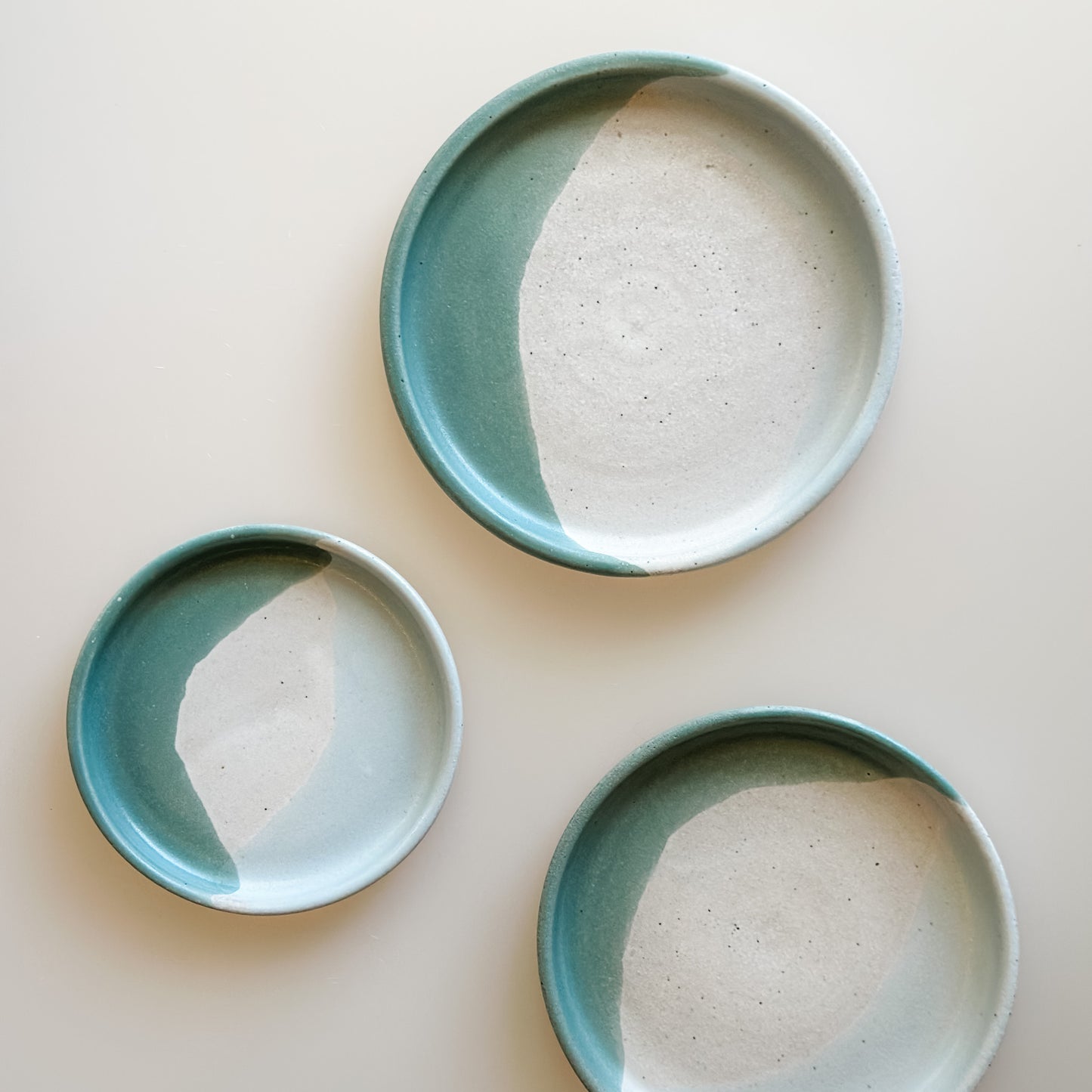 Three Tidal Handmade Ceramic Dishes sit on a cream background. They are glazed peacock-green, blue-grey, and white.