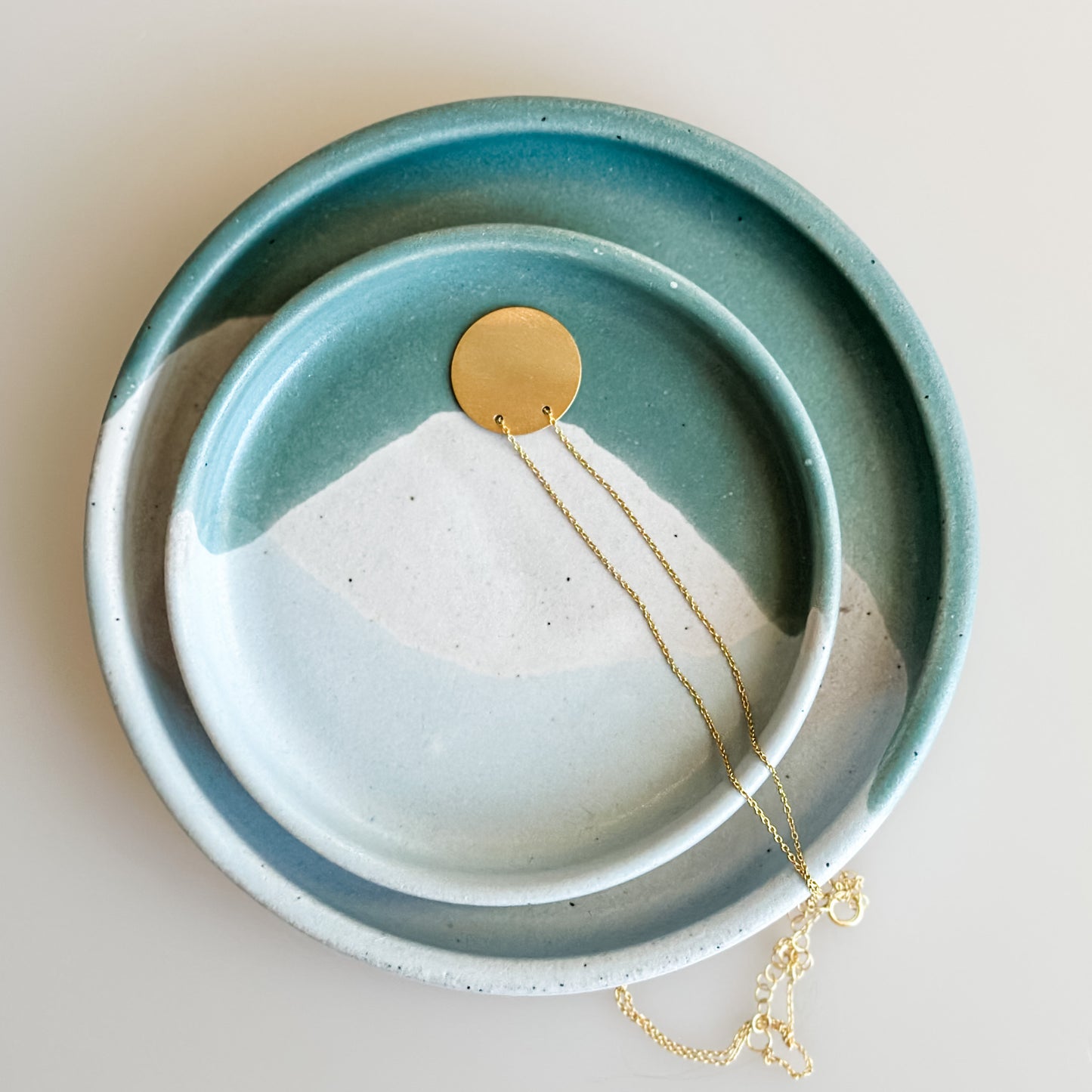 A small Tidal Handmade Ceramic dish sits inside a larger dish on a cream background. The dishes hold a gold necklace and are glazed peacock-green, blue-grey, and white.