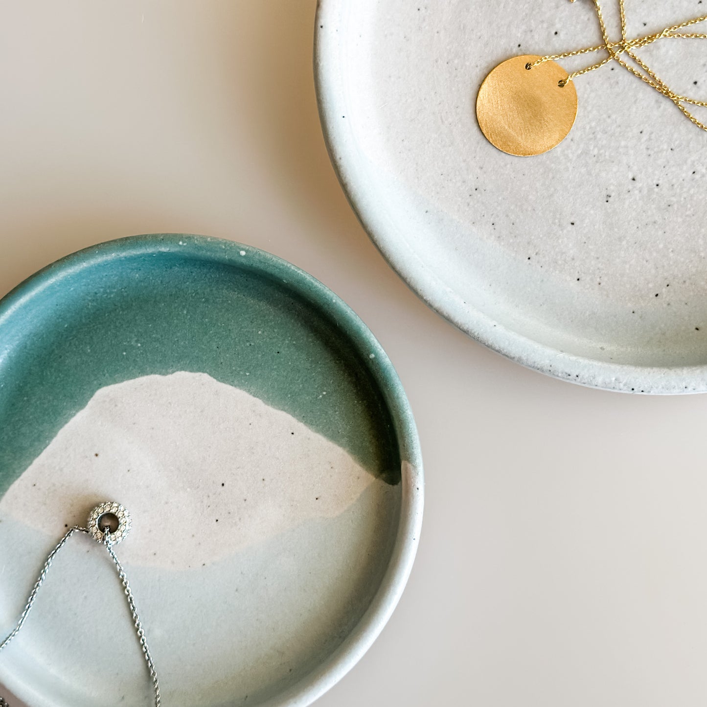 Two Tidal Handmade Ceramic dishes sit on a cream background. The dishes hold gold and silver necklaces and are glazed peacock-green, blue-grey, white and peach.