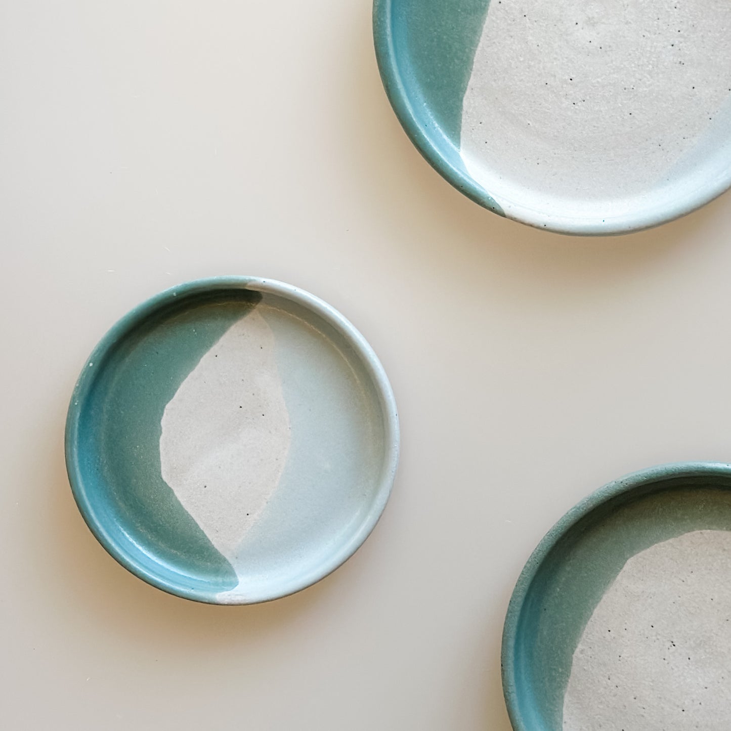 Three Tidal Handmade Ceramic dishes of different sizes sit on a cream background. They are glazed peacock-green, blue-grey, white and peach.