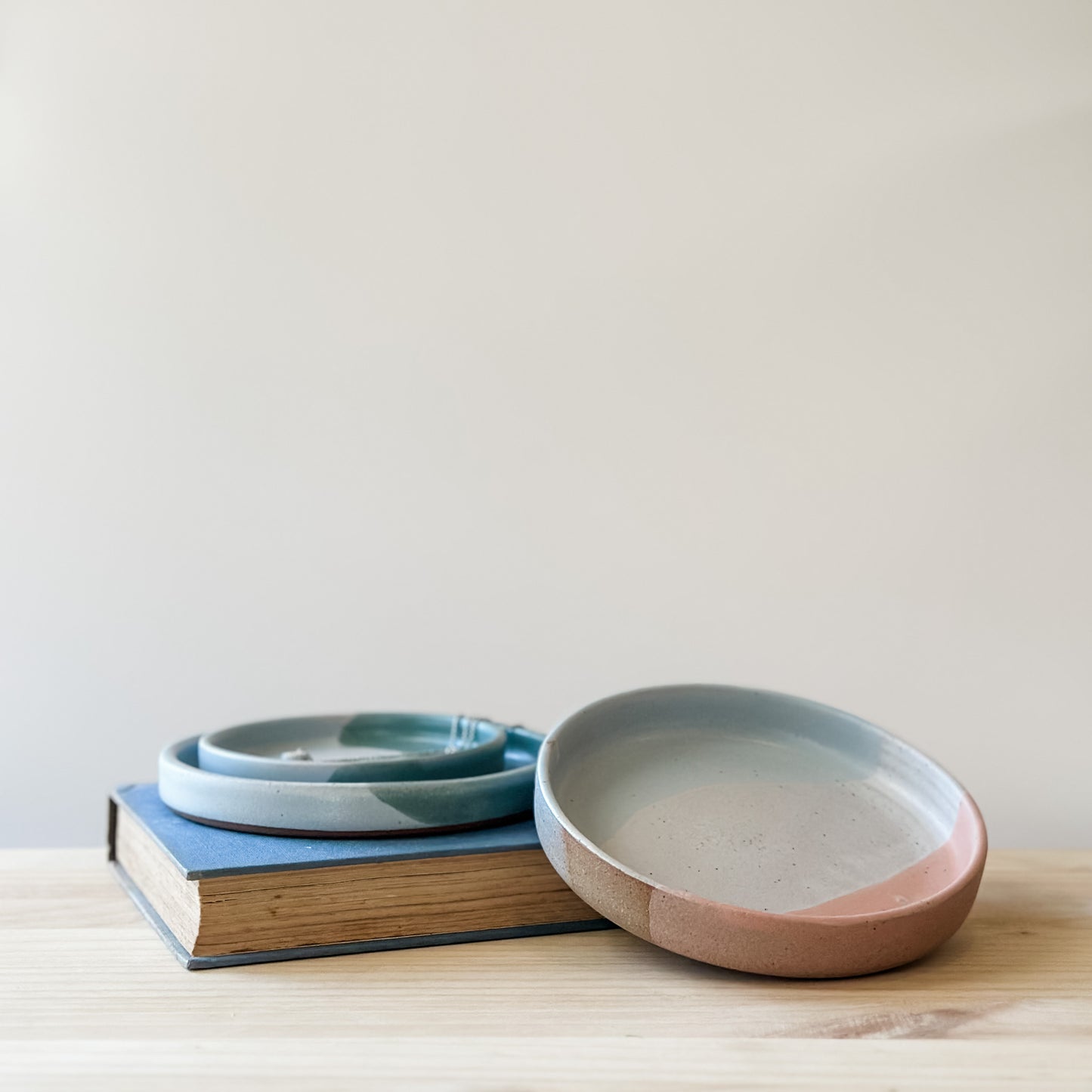Three Tidal Handmade Ceramic Dishes sit on a book and a wooden table. They are glazed peacock-green, blue-grey, white and peach.