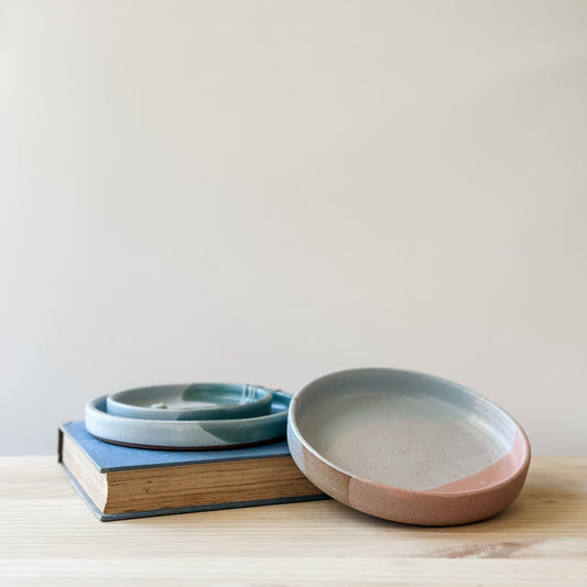 Three Tidal Handmade Ceramic Dishes sit on a book and a wooden table. They are glazed peacock-green, blue-grey, white and peach.