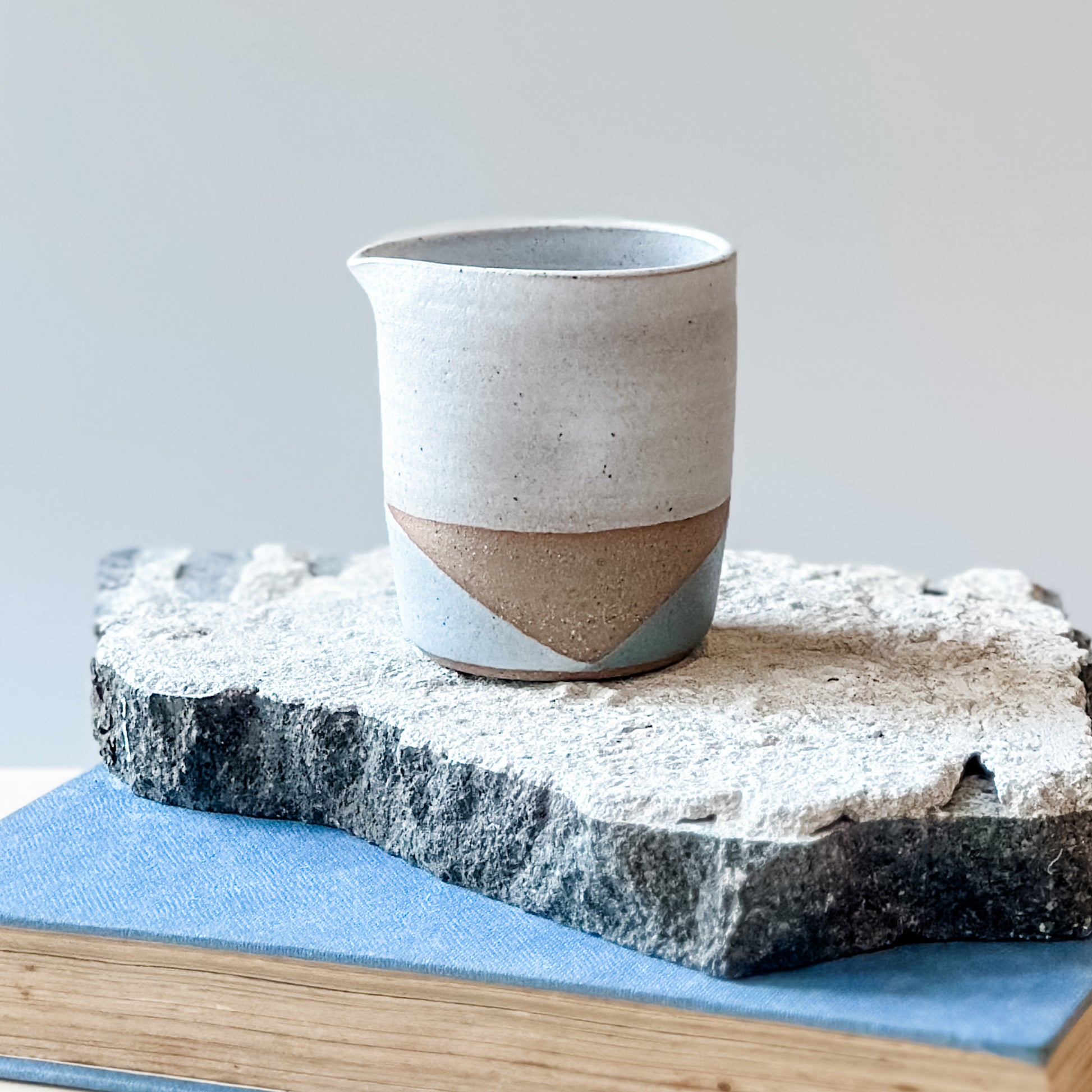 One creamer jug with white, grey and green glaze sits on a tile, a book and a wooden table.