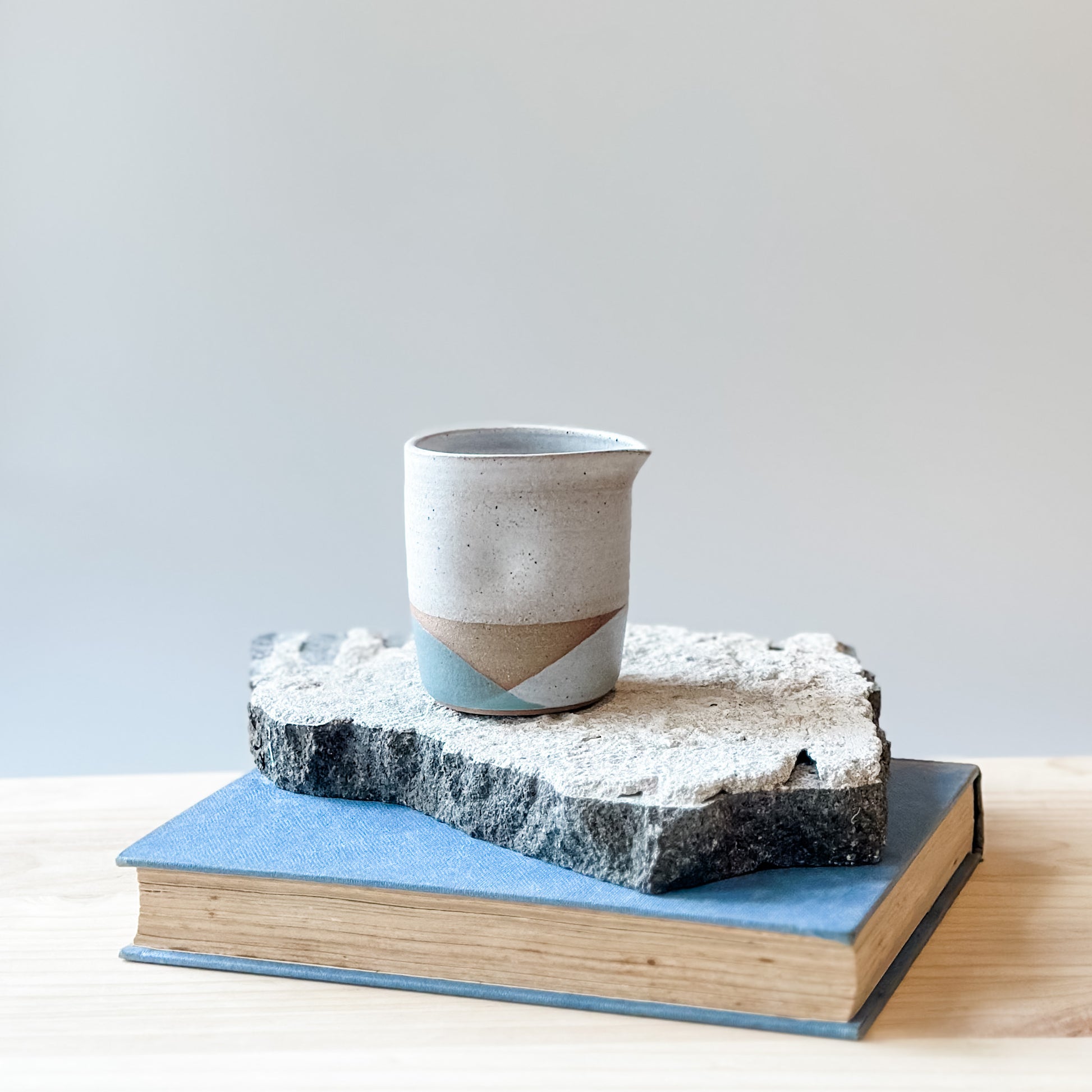 One creamer jug with white, grey and green glaze sits on a tile, a book and a wooden table.