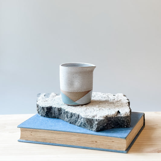 One creamer jug with white, grey and green glaze sits on a tile, a book and a wooden table.