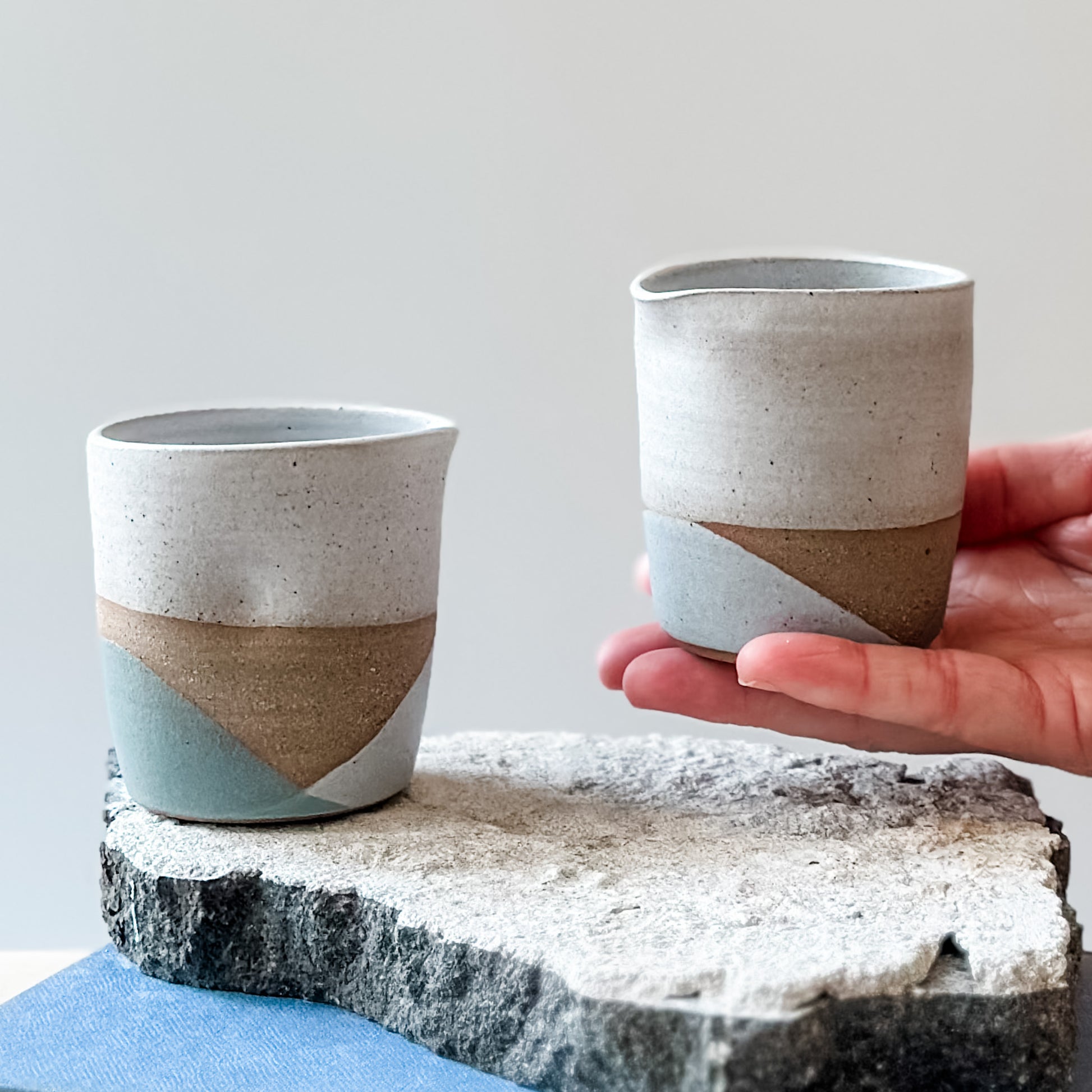 One creamer jug with white, grey and green glaze sits on a tile, a book and a wooden table. Another jug is held above the tile.