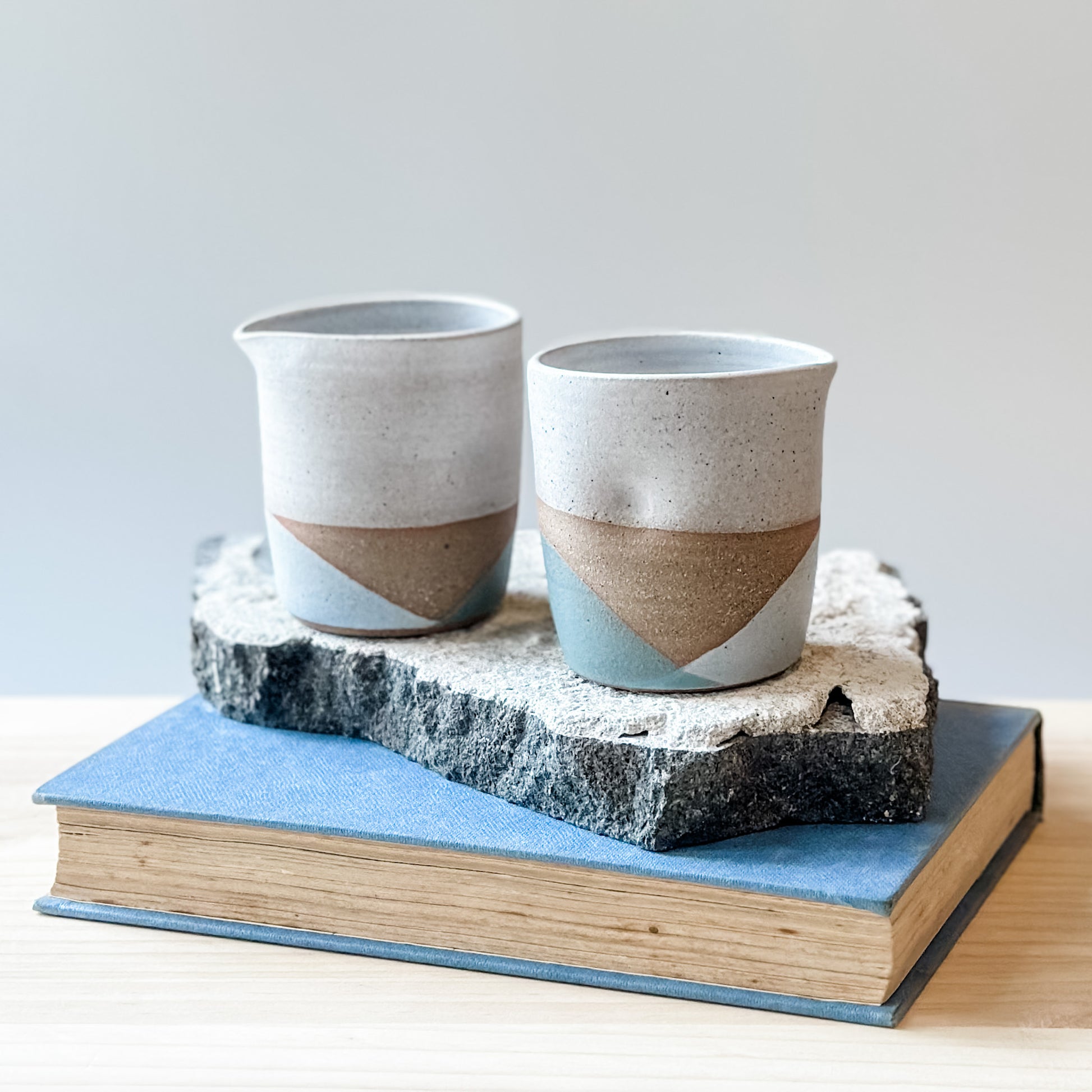 Two creamer jugs with white, grey, and green glaze sit on a tile, a book and a wooden table.