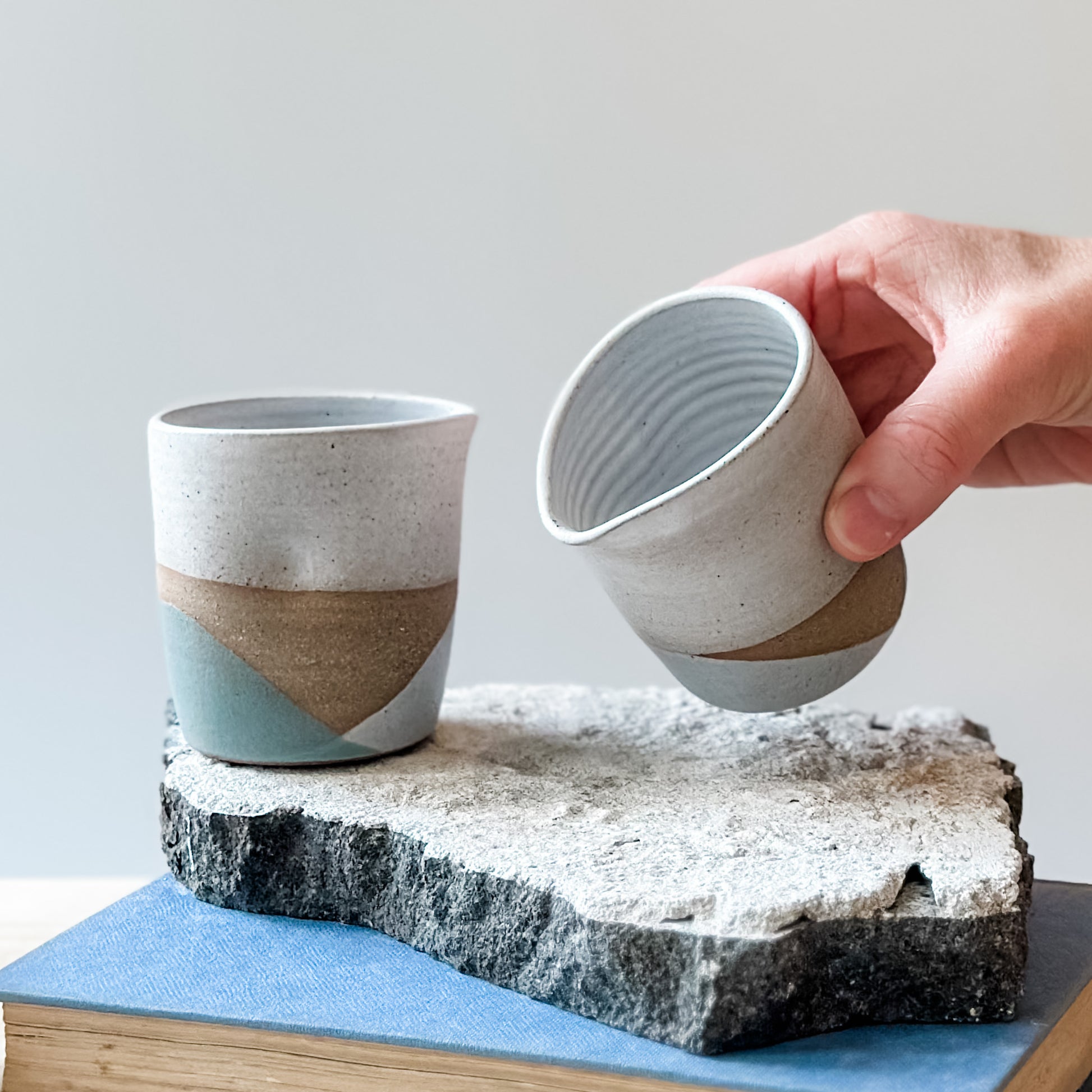 One creamer jug with white, grey and green glaze sits on a tile, a book and a wooden table. Another jug is held above the tile.