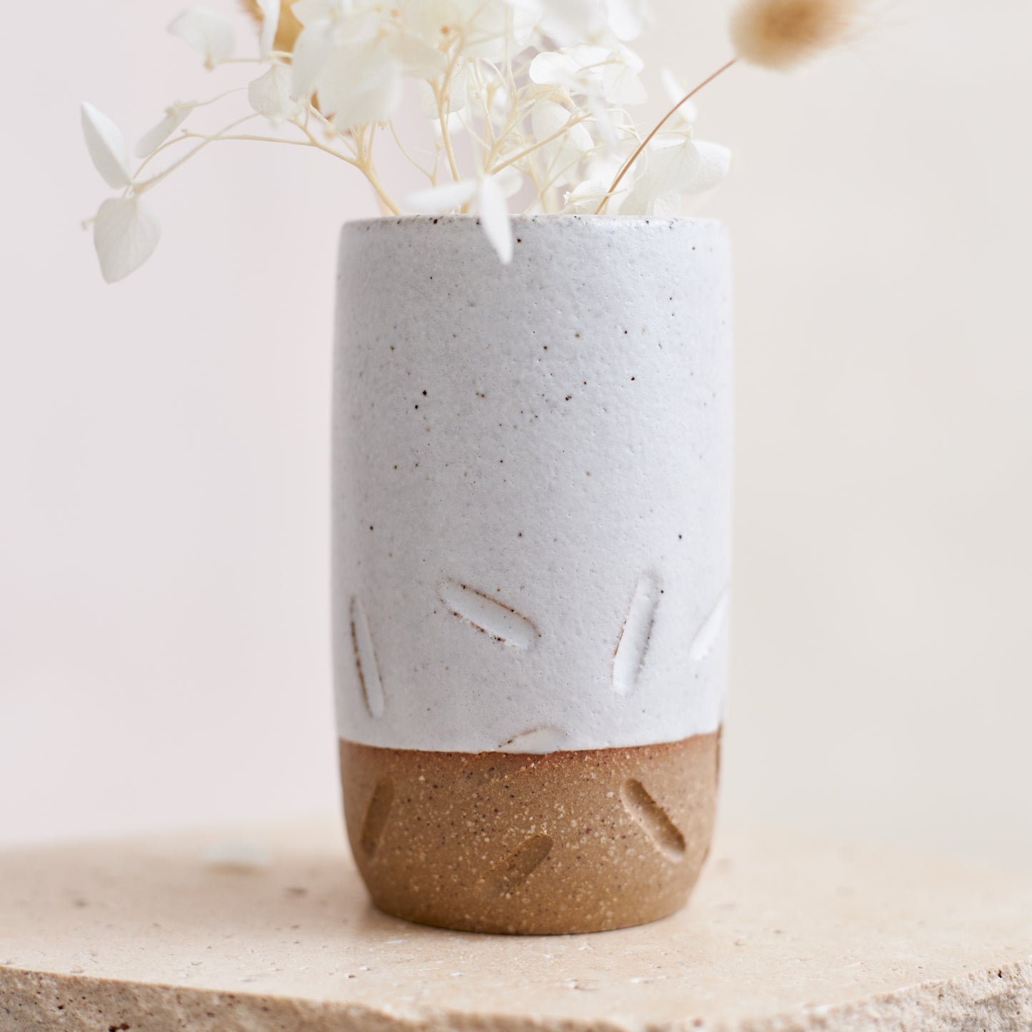 A close of the carving on the Warm Sands Handmade Ceramic Vase. The ceramic mini vase is holding dried coastal flowers. The vase is sitting on a cream coloured tile.