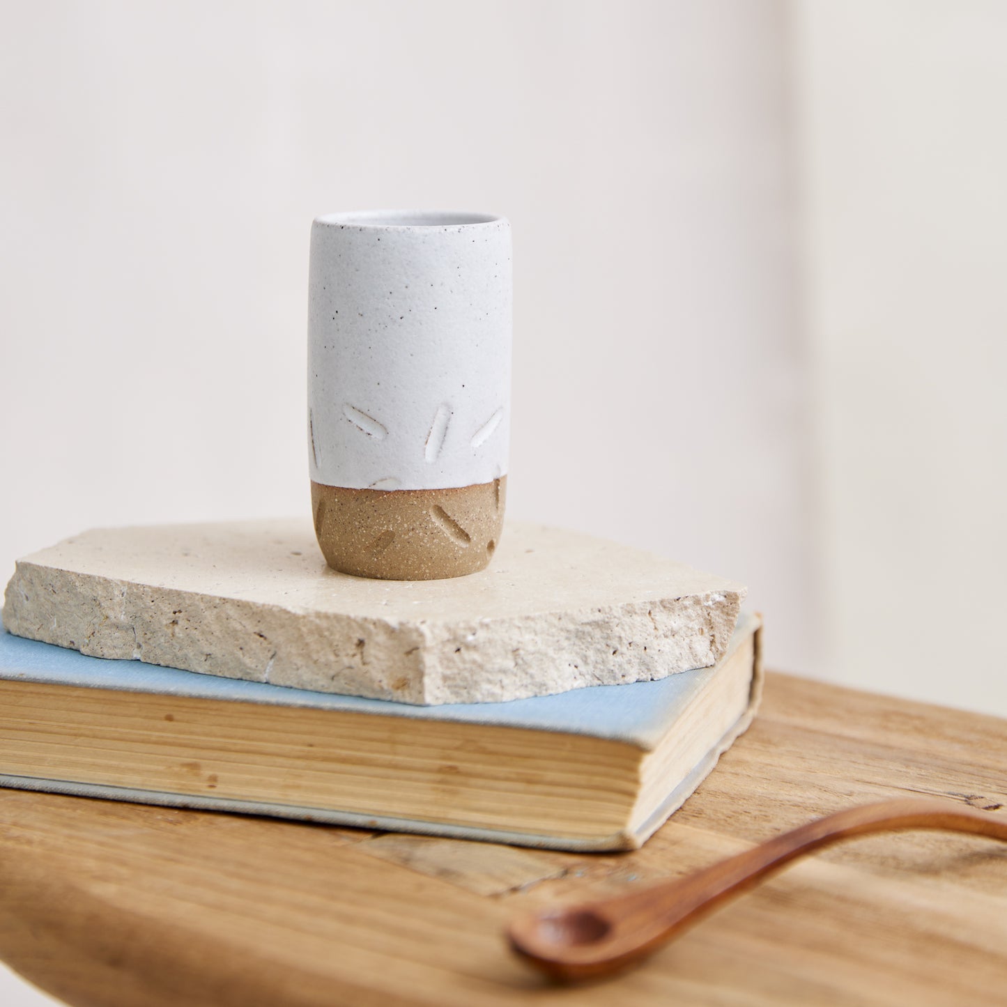 A cylindrical handmade ceramic vase in a white glaze. The cylindrical vase has carving detail on the bottom half. The handmade ceramic vase is sitting on a book and wooden side table in a coastal setting.