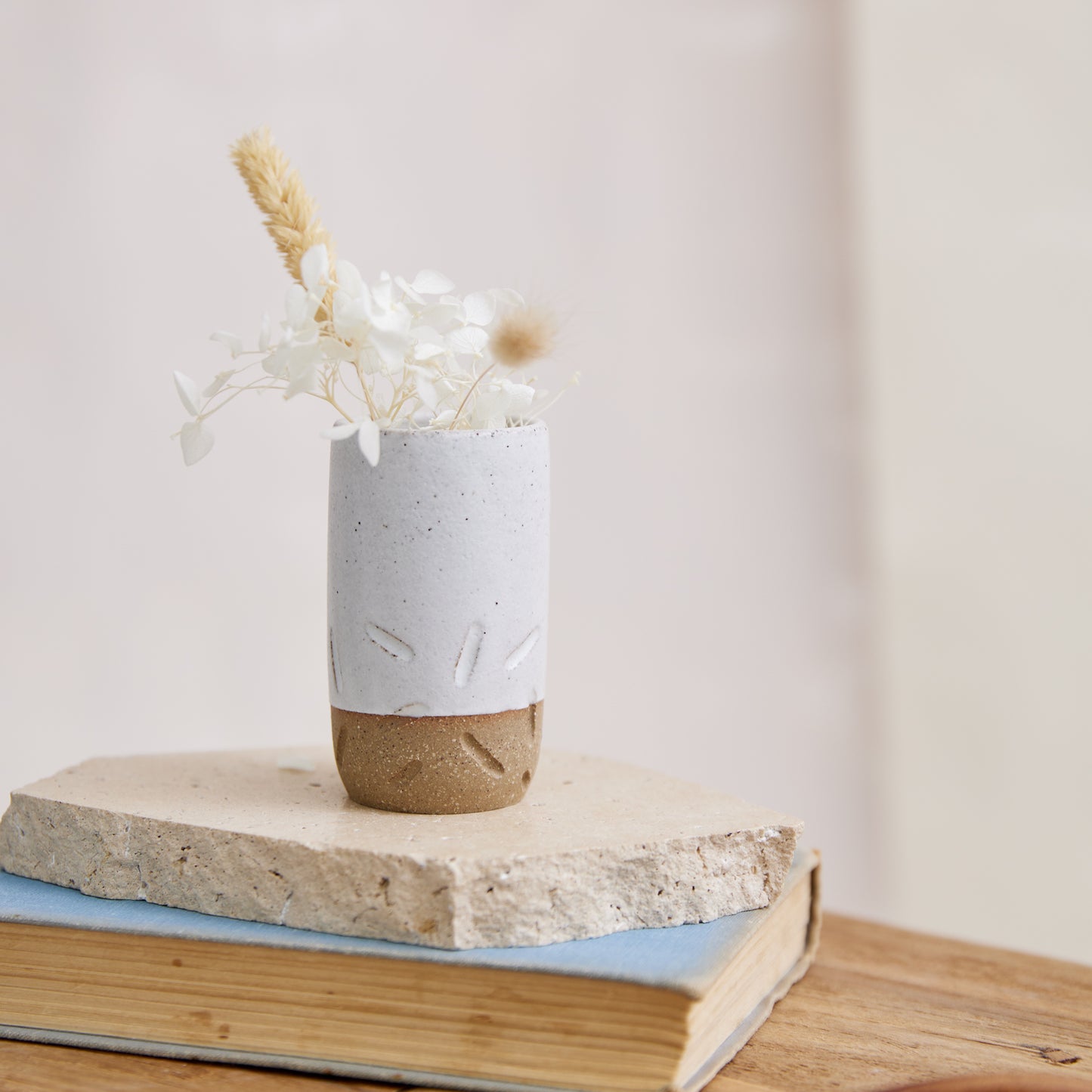 The Warm Sands Handmade Ceramic Vase is holding coastal flowers. This is a mini bud vase and has white glaze and carving detail on the bottom half. The handmade ceramic vase is sitting on a tile, book, and wooden side table.