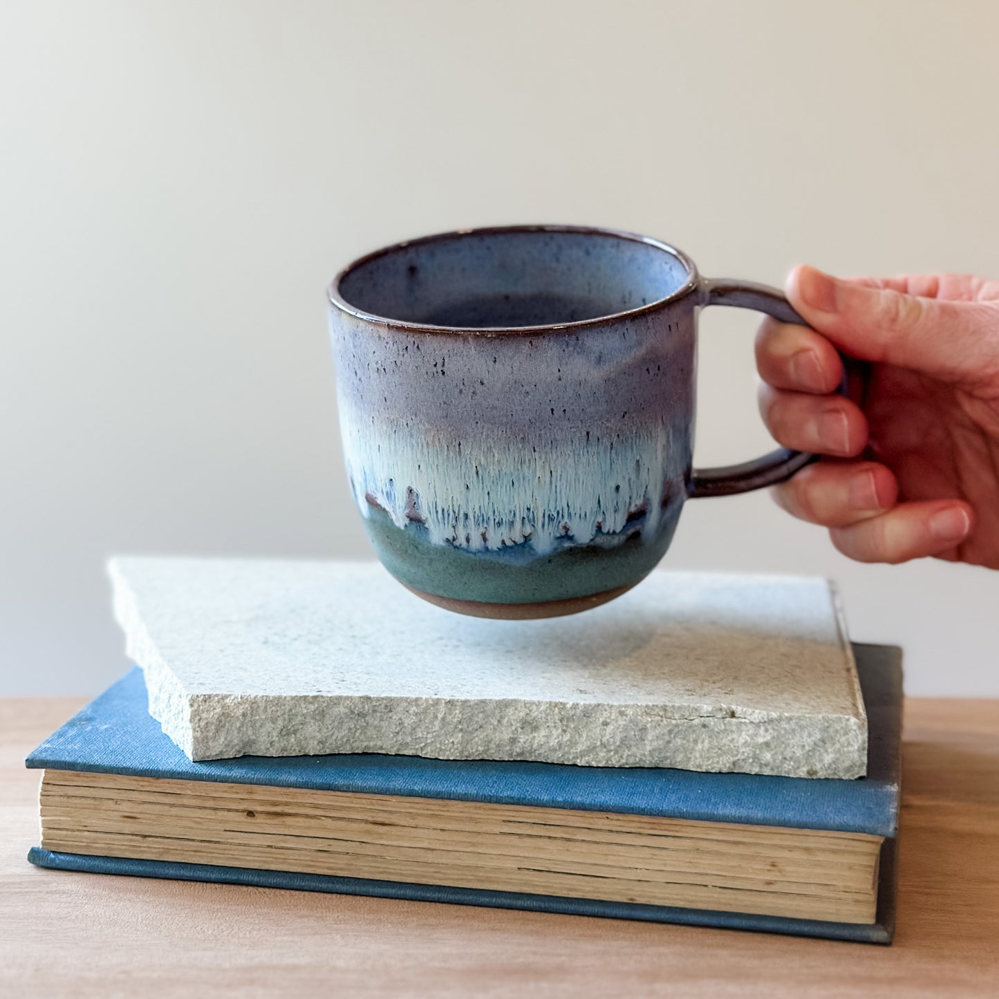 Hand holding a purple and blue ceramic mug over a stack of books on a wooden surface.