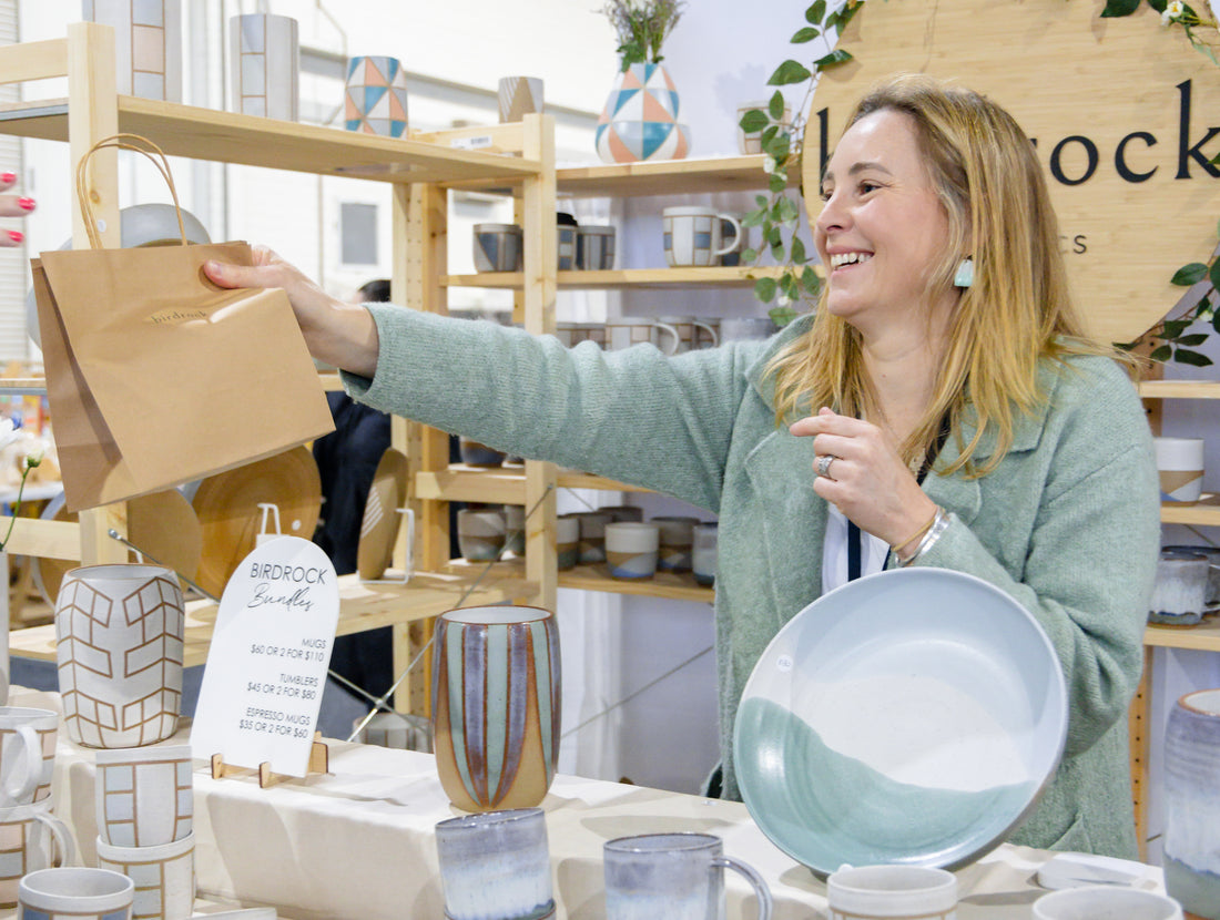 Melissa Tully passing over a branded Birdrock Ceramics bag to a customer. Melissa is standing in her market stall of handmade ceramics, displaying ceramic vases, plates, bowls mugs, and tumblers.